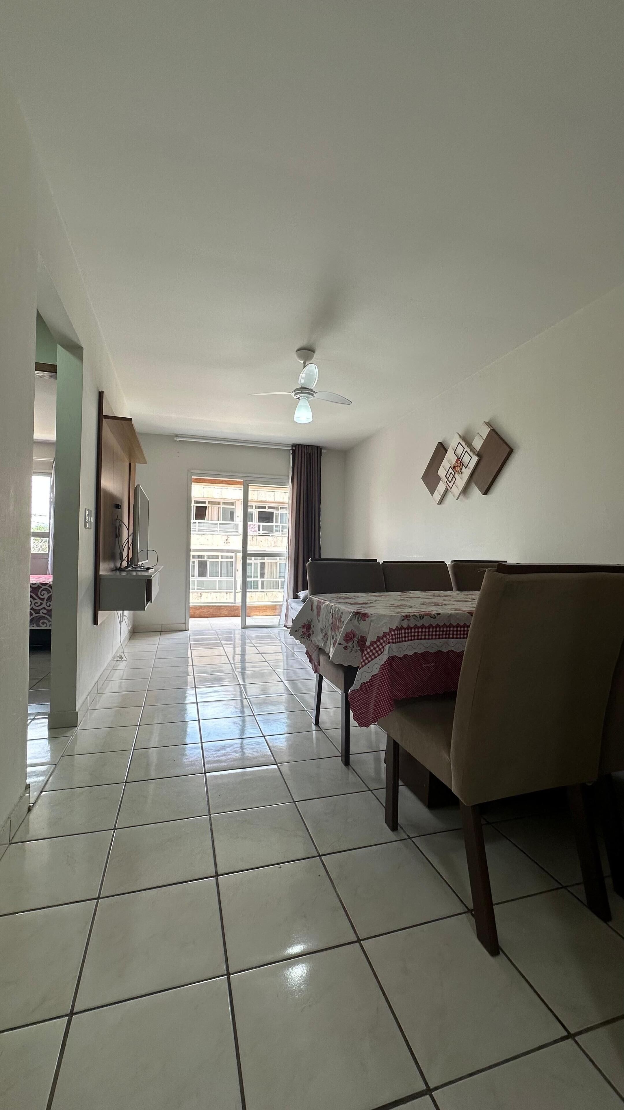 Una habitación amplia con piso de baldosas, un ventilador de techo, una mesa de comedor con sillas y vista a un edificio por una puerta de vidrio.