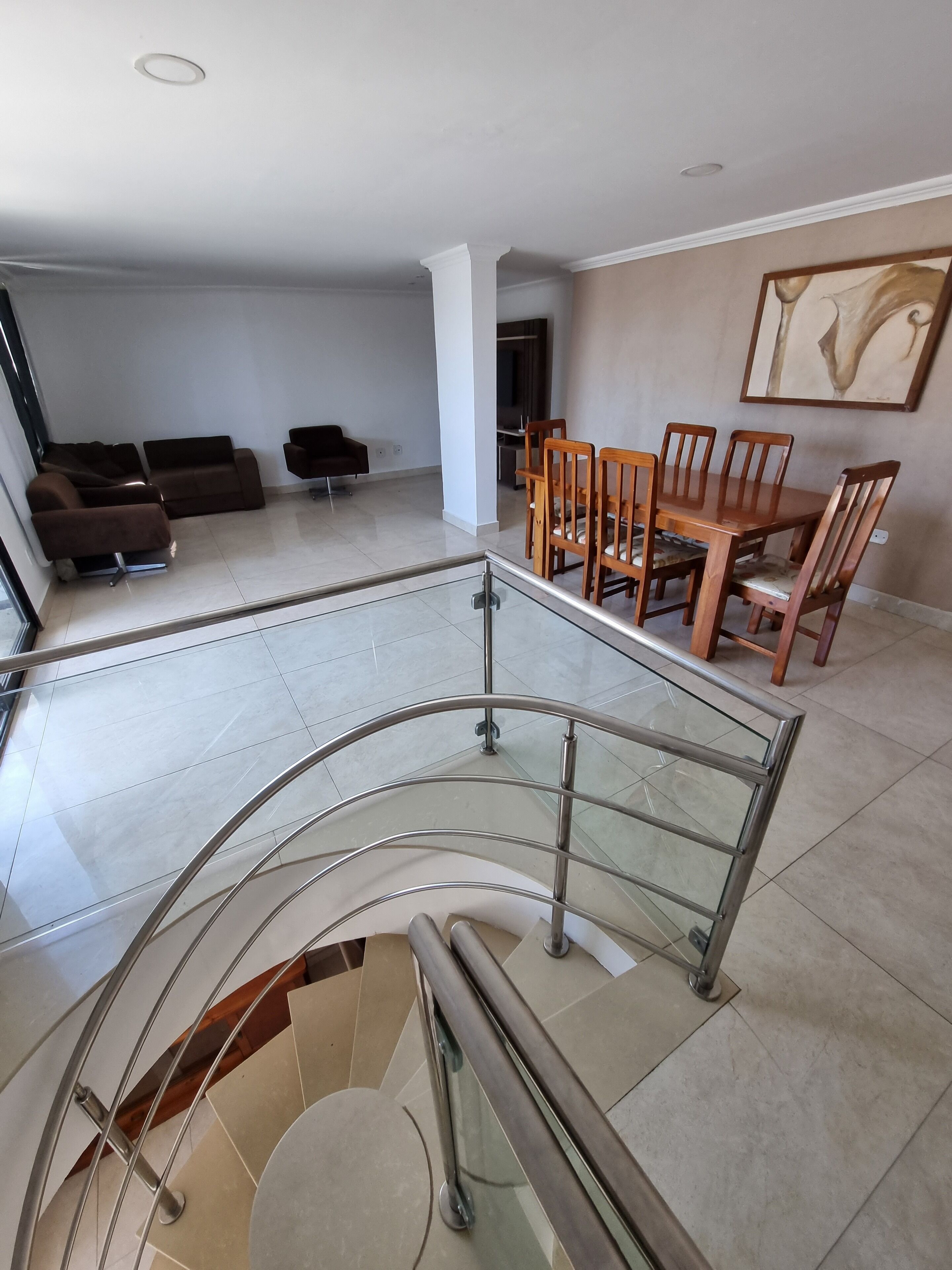 A spacious living area with a dining set and sofas, a staircase with glass railings, and a modern interior design.