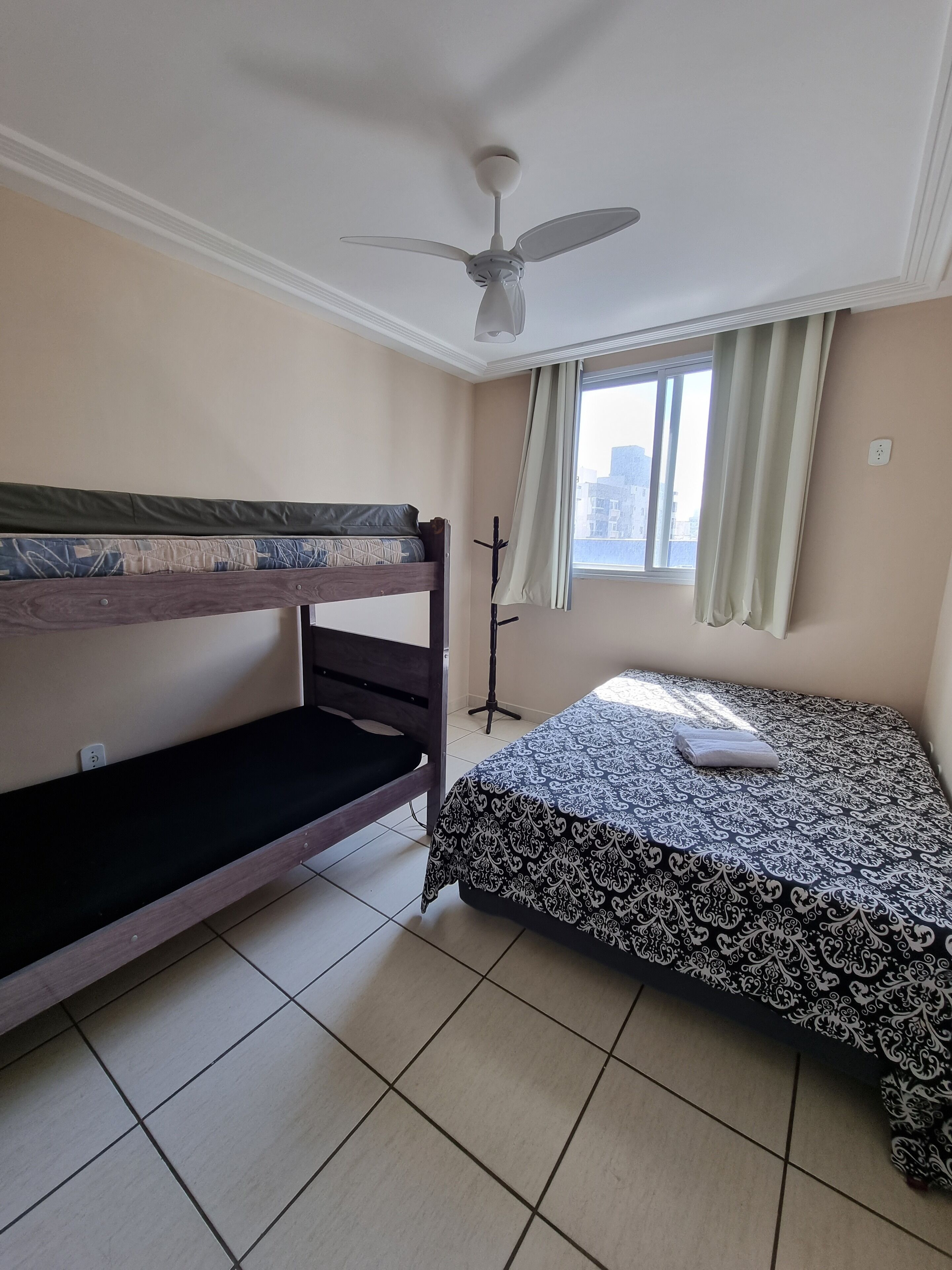 A room with a bunk bed, a single bed with a patterned bedspread, a standing lamp, and a window with curtains.