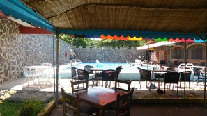 Un espace piscine agrémenté d’un mur en pierre, un coin salon couvert avec des tables et des chaises, et un toit de chaume.