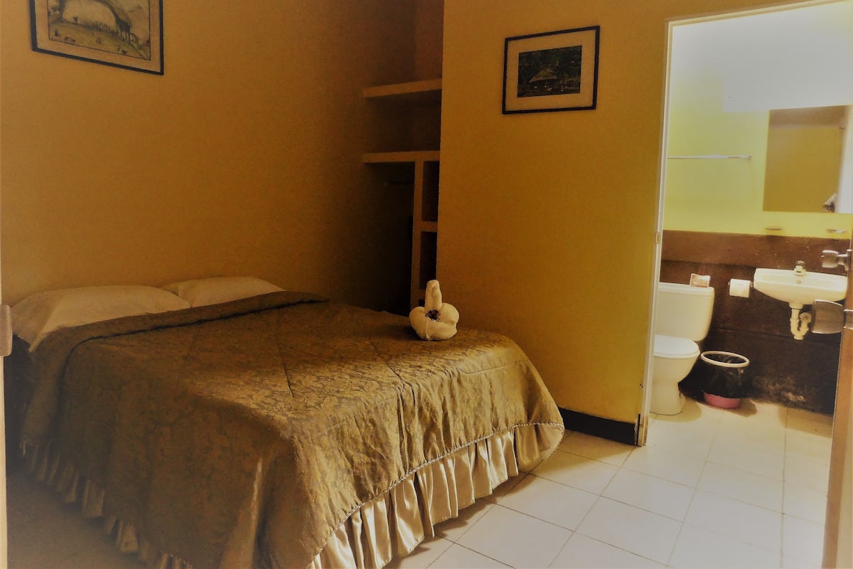 A bedroom with a bed, a nightstand, a framed picture on the wall, and a bathroom visible through an open door.