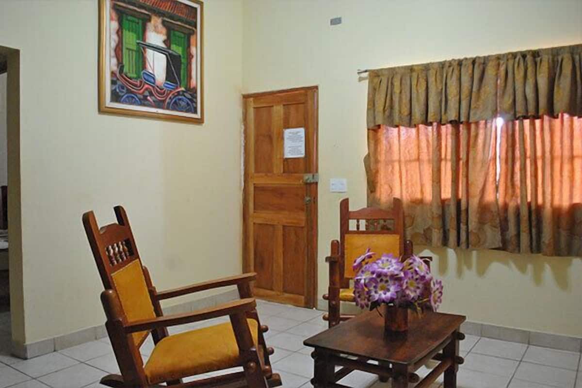 A room with a wooden chair, a small table with a vase of flowers, a door, and a painting on the wall.