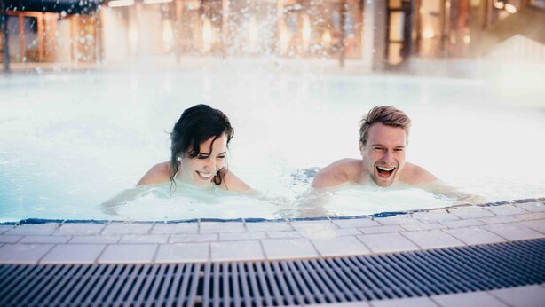 Ett par i ett bubbelbad med ett kaklat golv, och en byggnad syns i bakgrunden.