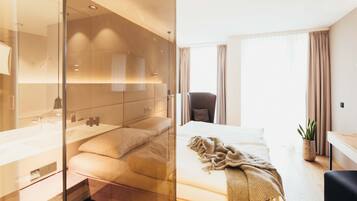 A modern hotel room with a glass partition separating the bathroom and the bedroom. The bathroom features a large mirror, a sink, and a vanity. The bedroom has a bed with a blanket, a chair, and a potted plant.
