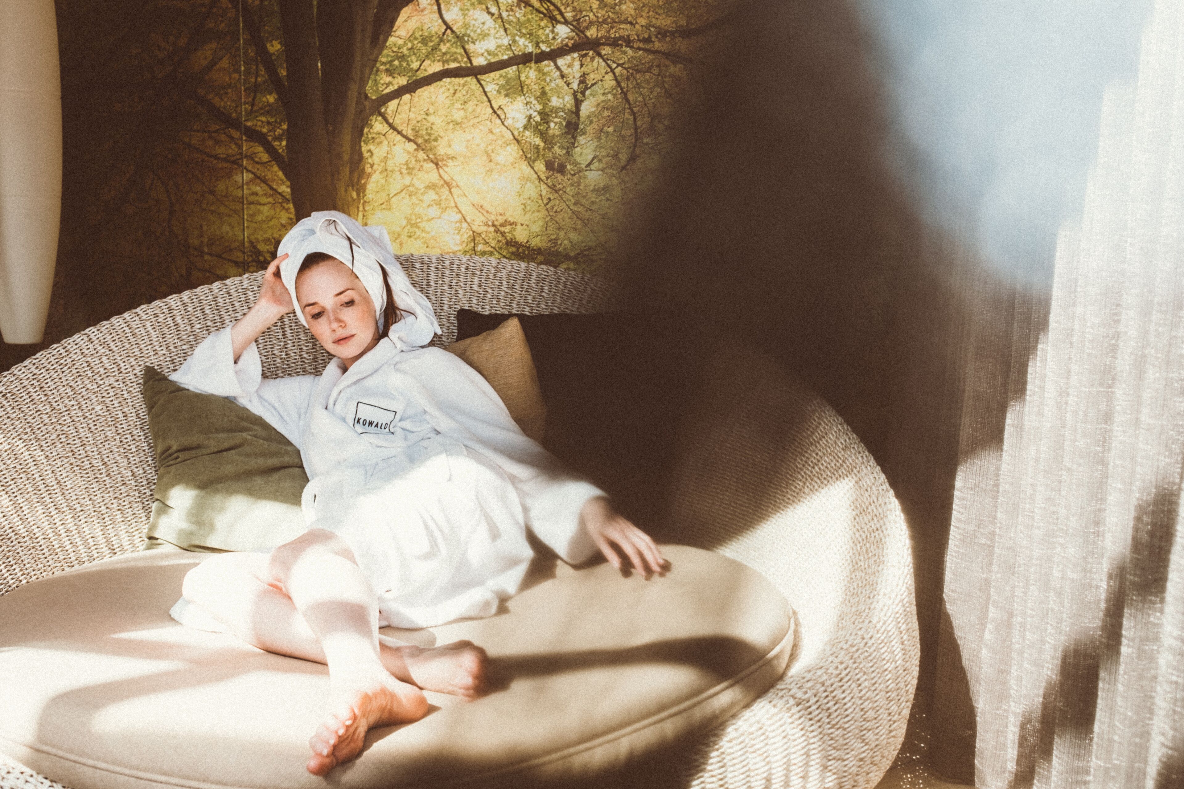 A child sitting on a wicker sofa with a view of trees through a window.