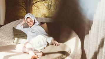 A child sitting on a wicker sofa with a view of trees through a window.