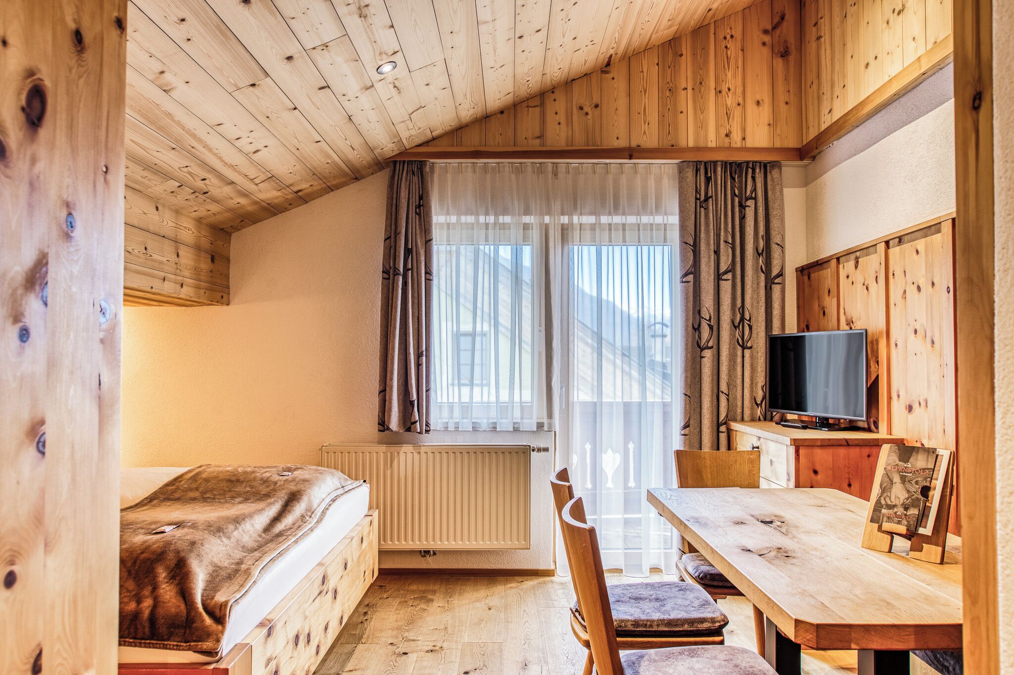 A room with a bed, a wooden table, a television, and a window with curtains.