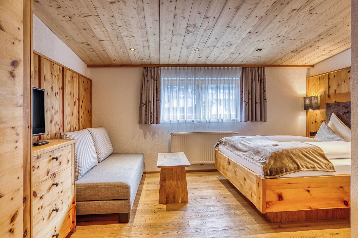 A room with a wooden ceiling, a bed, a sofa, a TV, and a window with curtains.