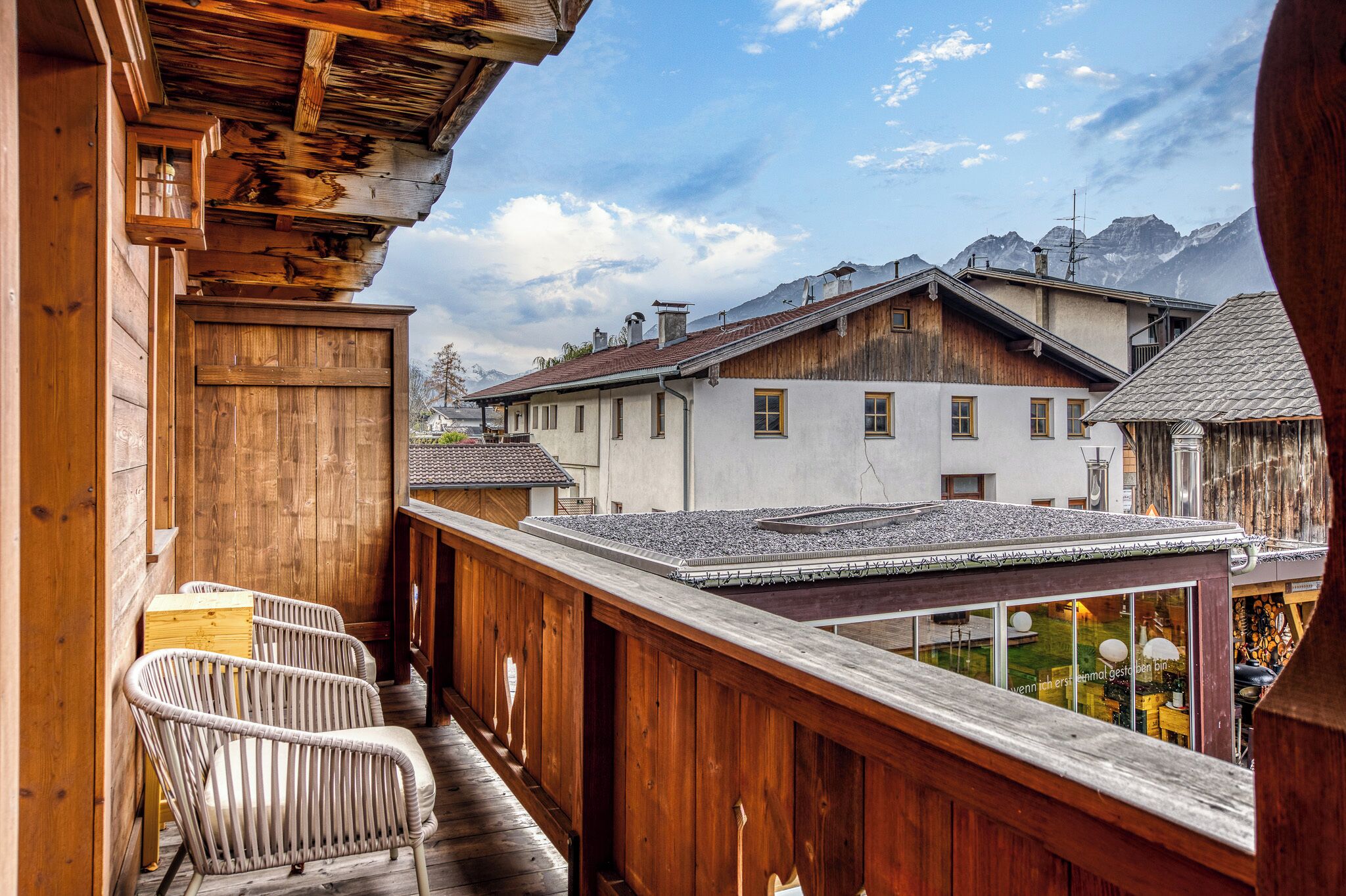 Un balcón de madera con sillas de mimbre, vista a edificios tradicionales y montañas al fondo.