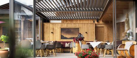 Een modern terras met houten tafels en stoelen, een glazen overkapping en een houten wand met een schilderij.