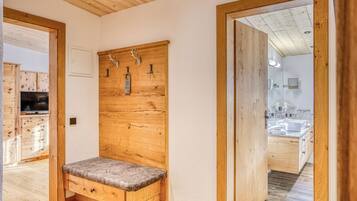Un banc en bois avec un siège rembourré, un mur en bois muni de crochets et une porte en bois donnant accès à une salle de bain.