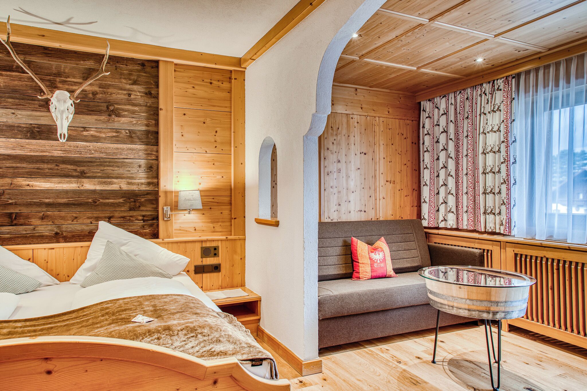 A wooden interior with a bed, a sofa, a glass table, and a wall-mounted deer skull.