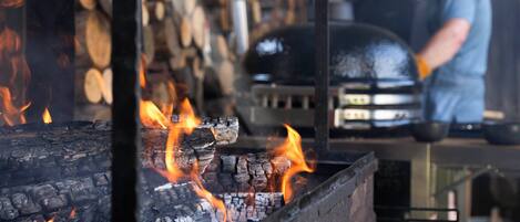 A grill with burning wood and flames, and a person cooking in the background.