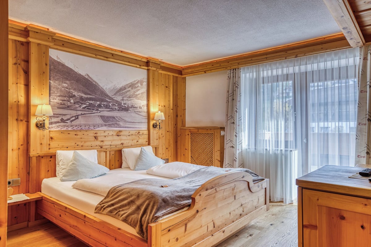 A wooden room with a bed, a desk, and a large window with curtains.