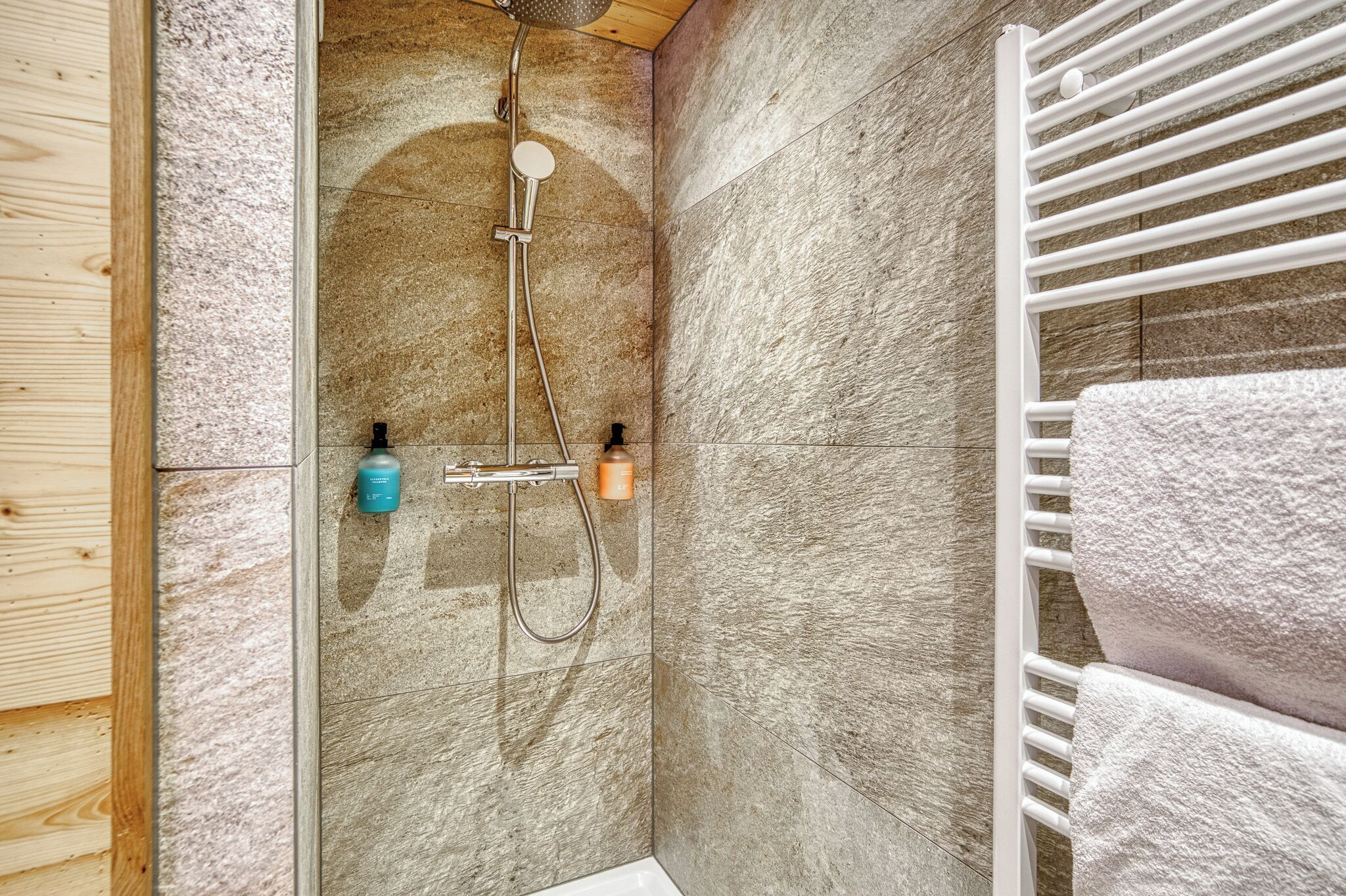 Una ducha con cabezal de ducha manual y grifo empotrado, un perchero con una toalla blanca y un panel de madera a la izquierda.