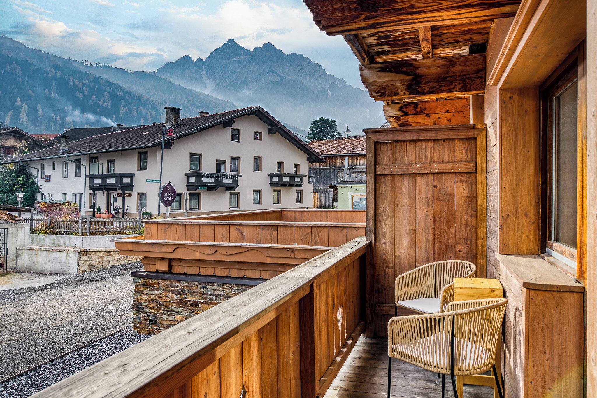 A wooden balcony with wicker chairs, a stone wall, and a view of a mountain.