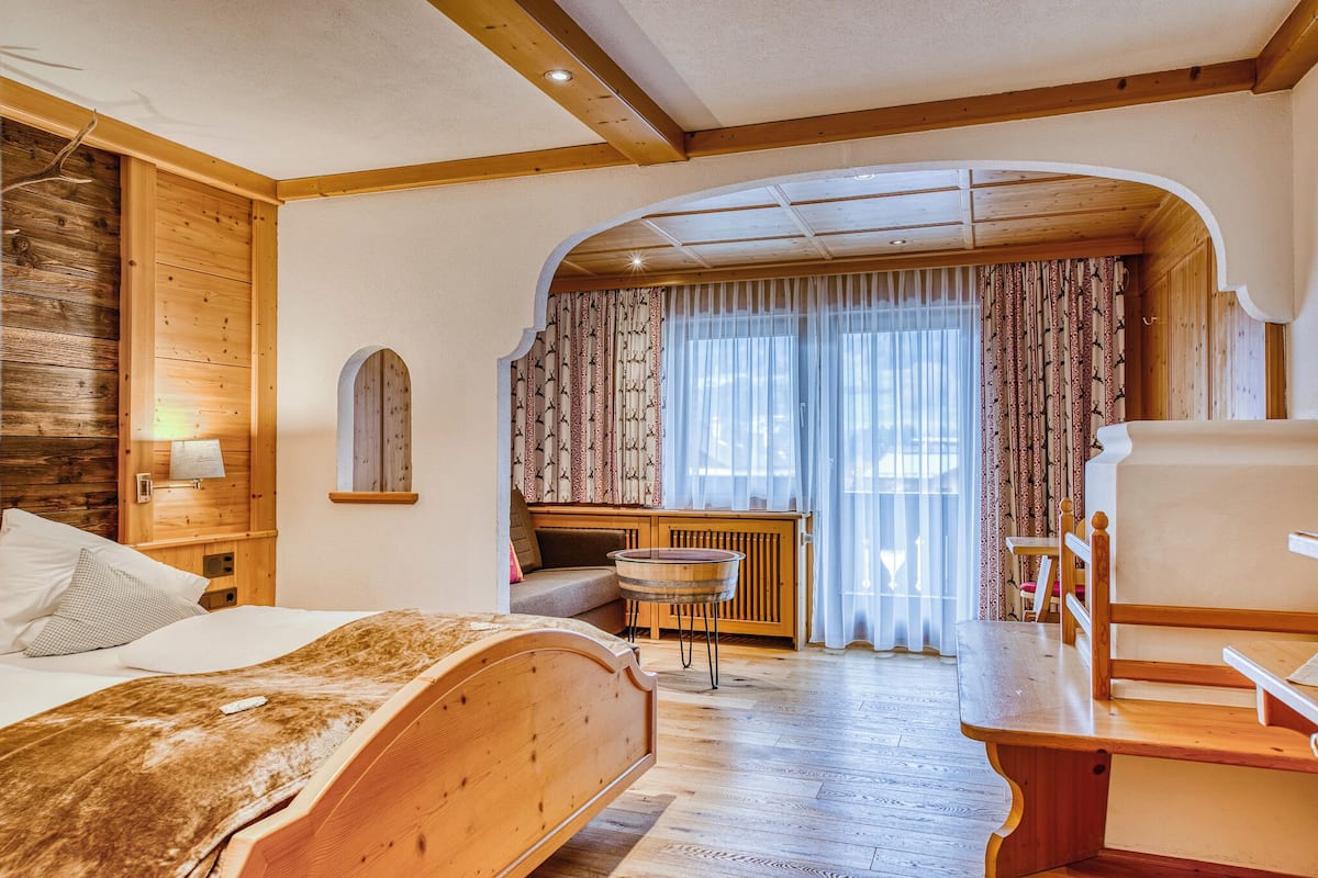 A room with a bed, a wooden bench, a small table, a bench with a backrest, and a window with curtains.