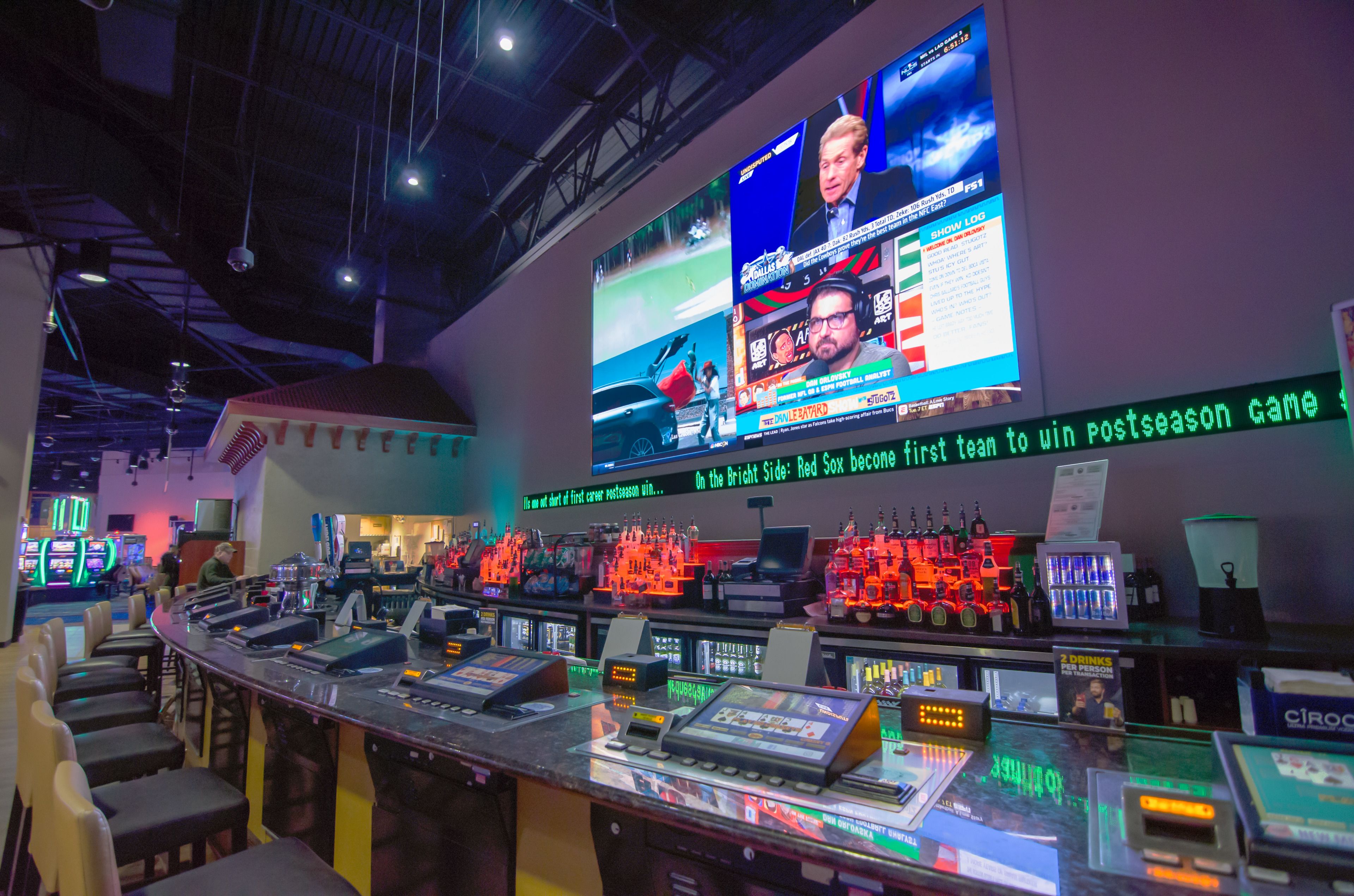 A bar area with multiple gaming machines and a large screen displaying sports content.