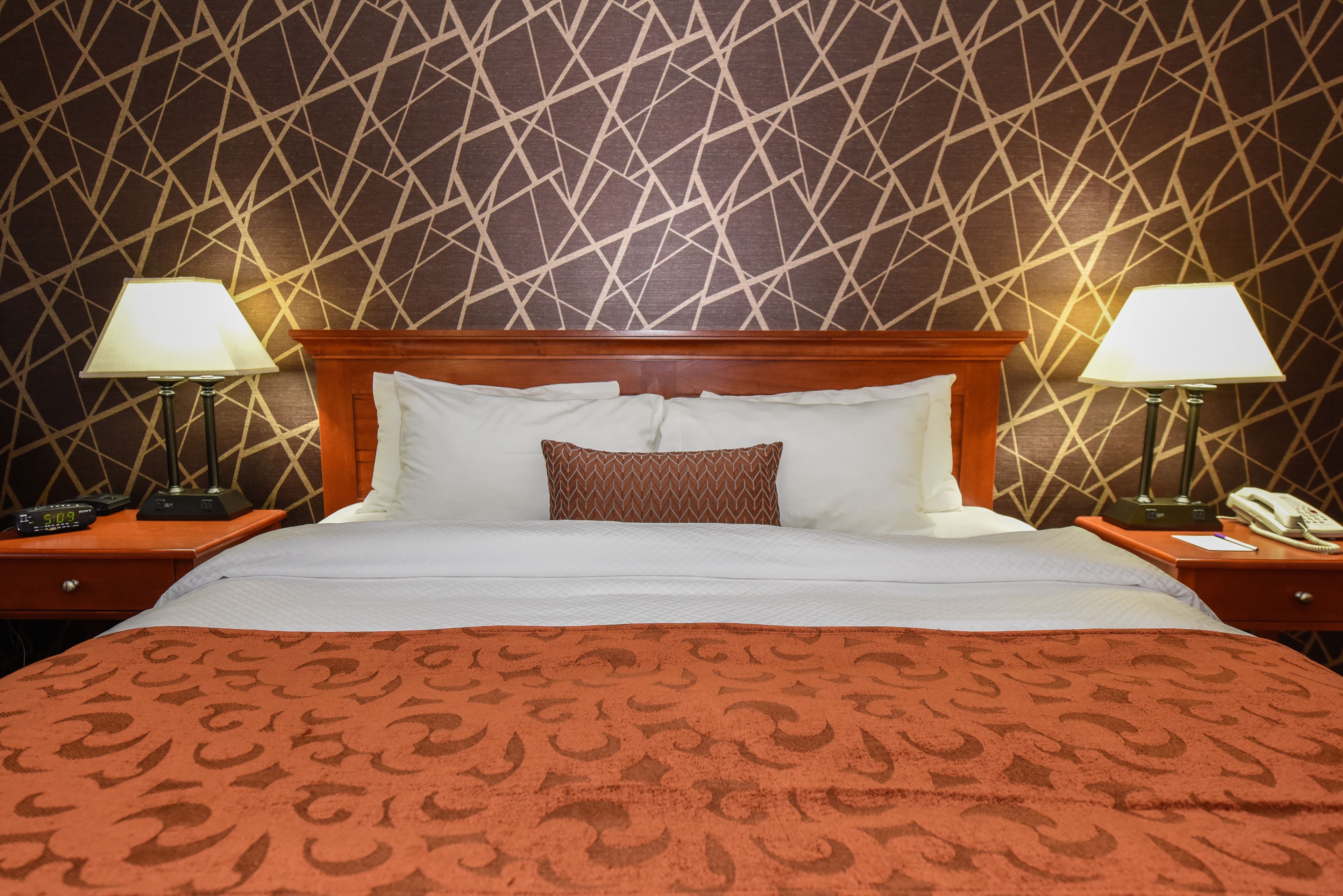 Una habitación de hotel con papel tapiz estampado, cabecero de madera, cama con ropa de cama blanca y marrón, y dos mesitas de noche con lámparas y teléfonos.