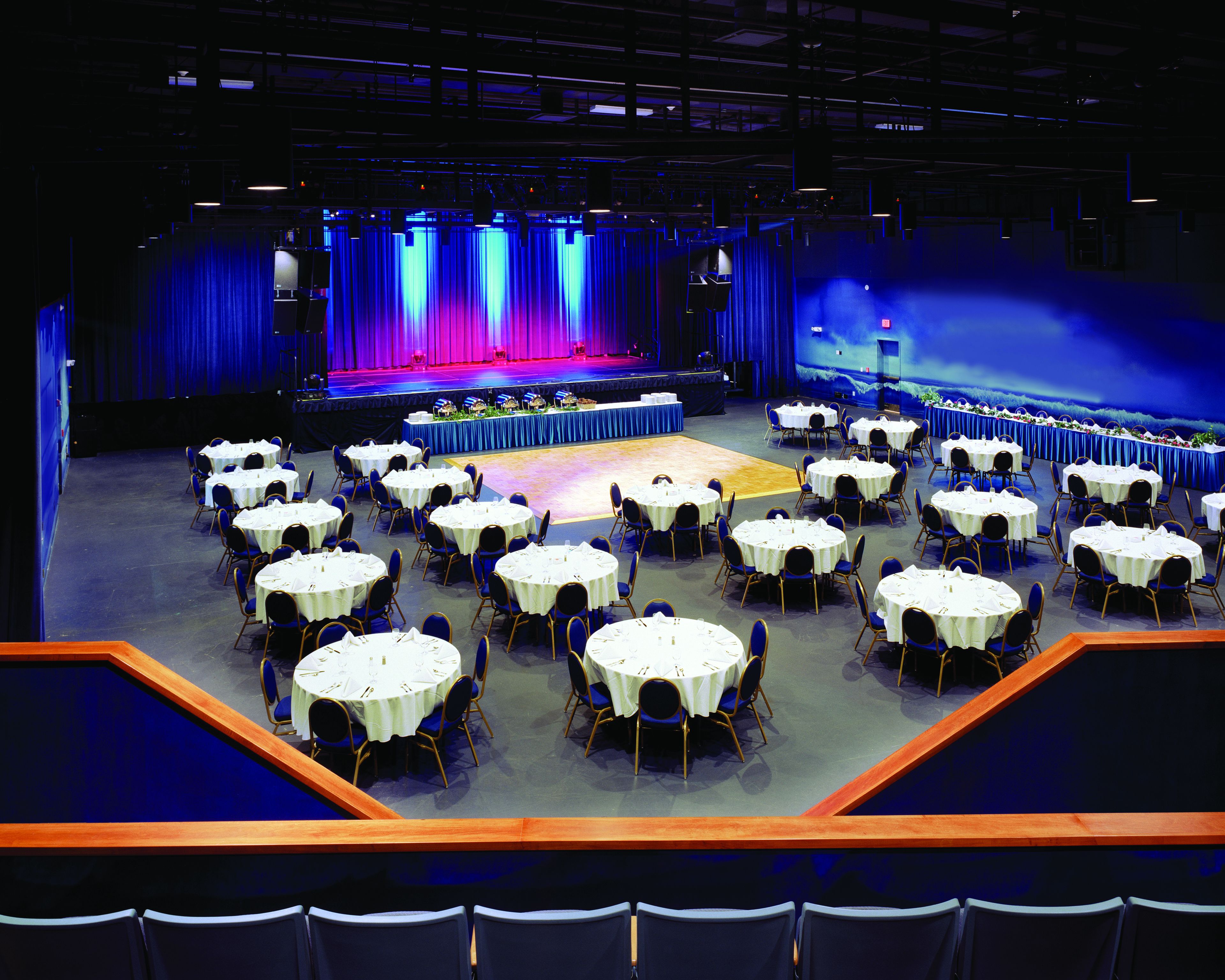 A large hall with round tables and chairs, a stage with a blue curtain backdrop, and a seating area with a view of the stage.