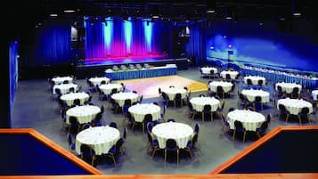 Een ruime zaal met ronde tafels en stoelen, een podium met een blauw gordijn als achtergrond, en een zitgedeelte met uitzicht op het podium.