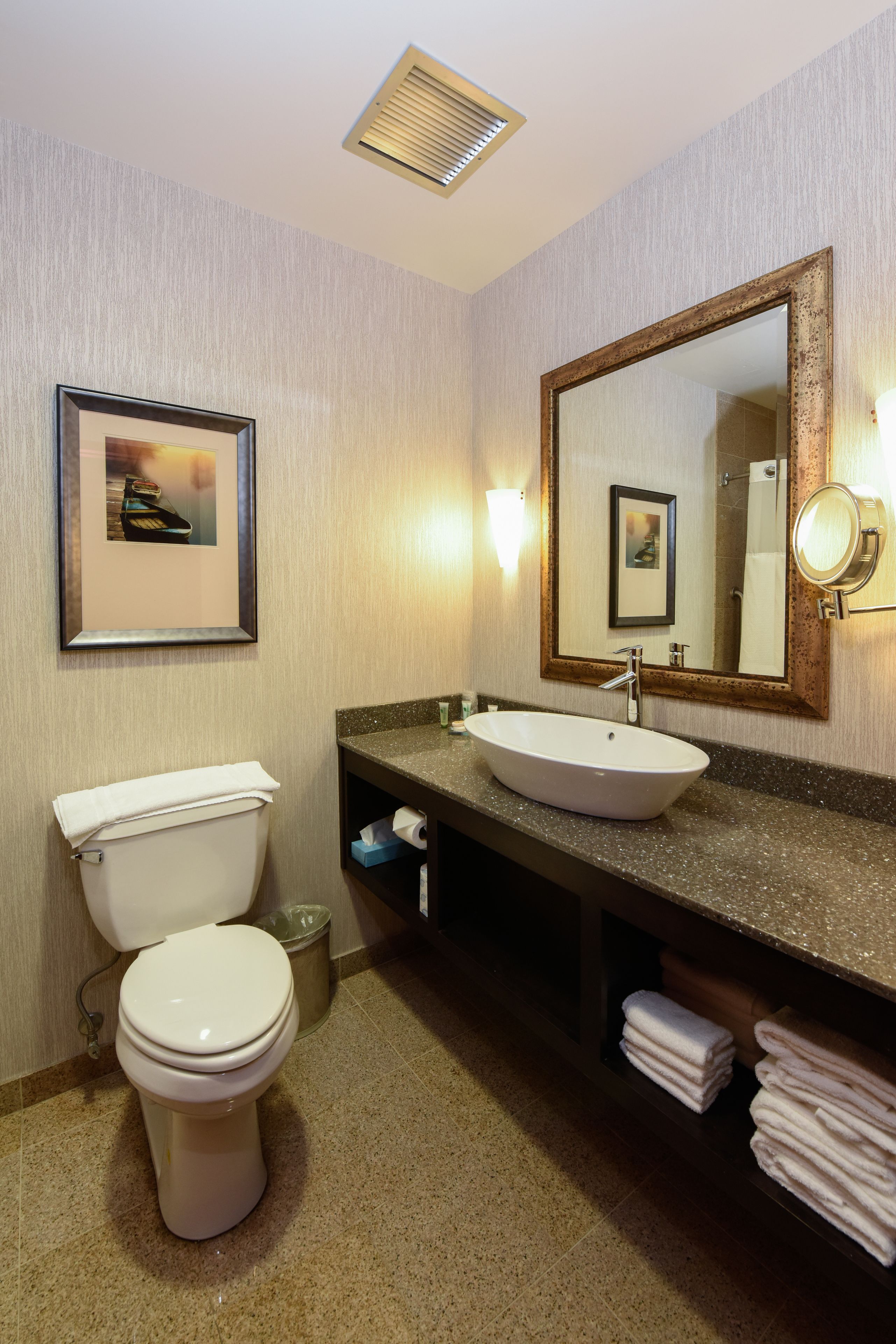A hotel bathroom with a toilet, sink, and mirror. There is a framed picture on the wall and a towel rack with towels.