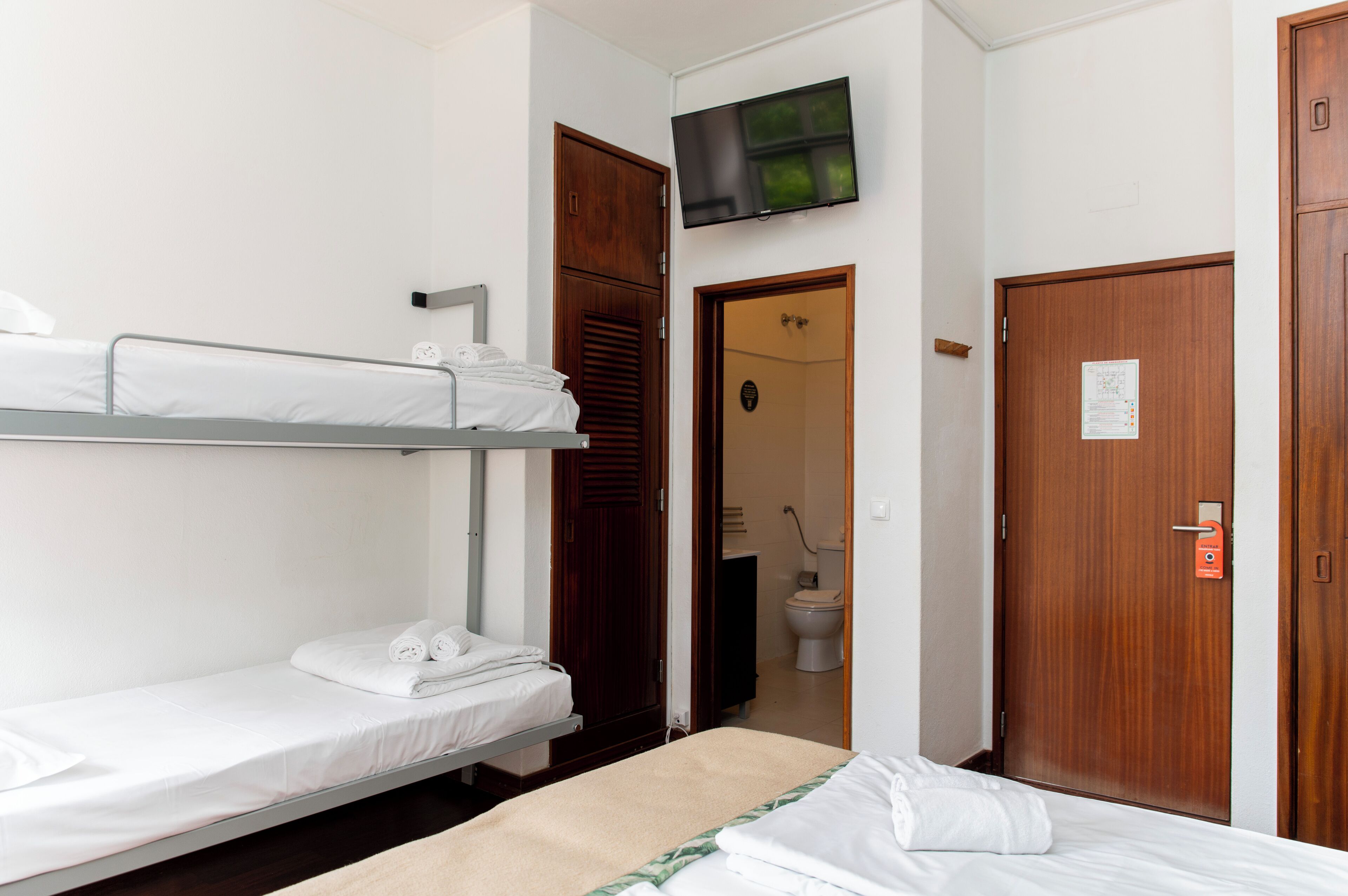 A hotel room with a bunk bed, a single bed, a television mounted on the wall, and a wooden door.