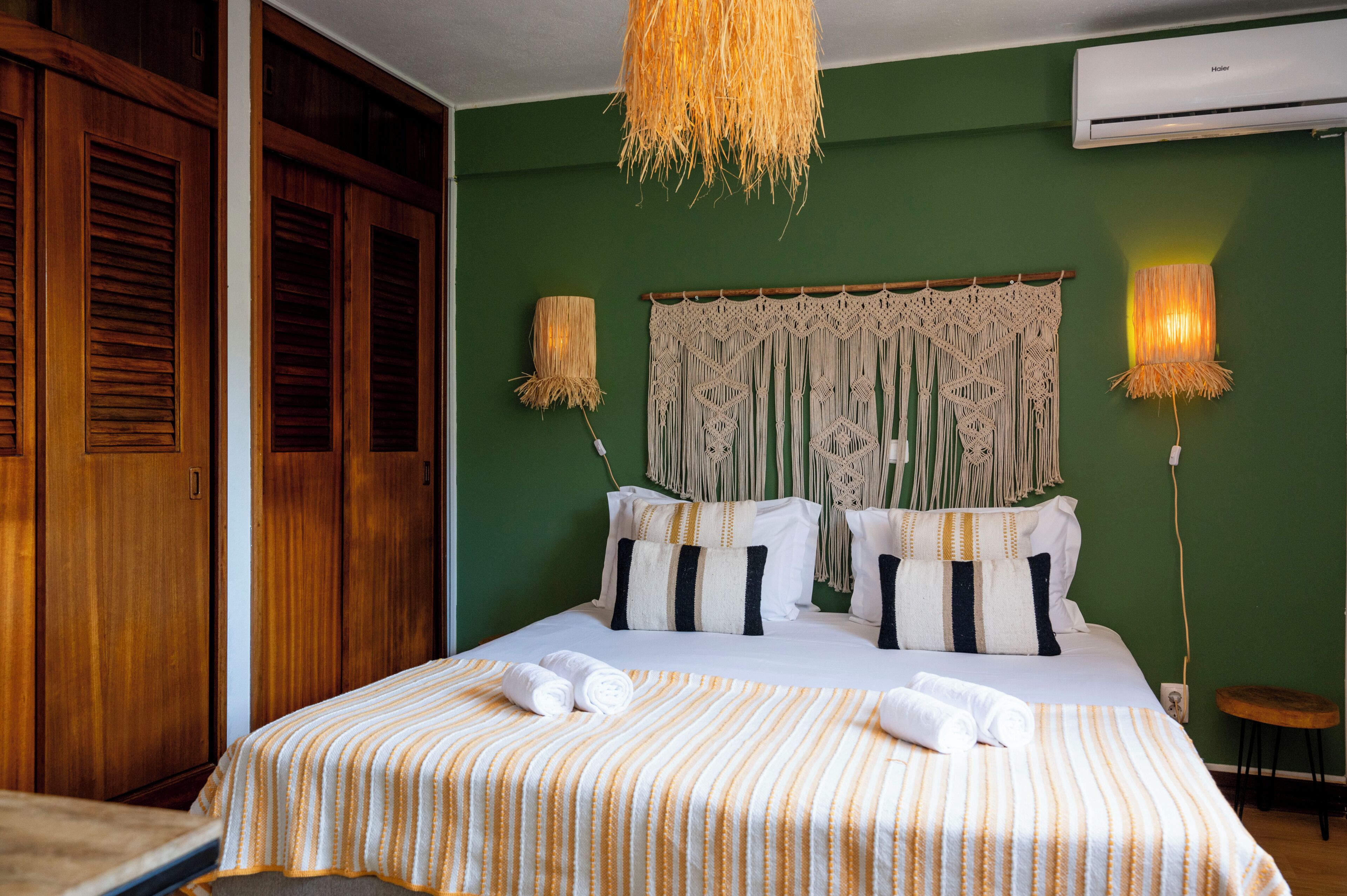 A hotel room with a bed, wooden wardrobe, macramé wall hanging, and two wall-mounted lamps.