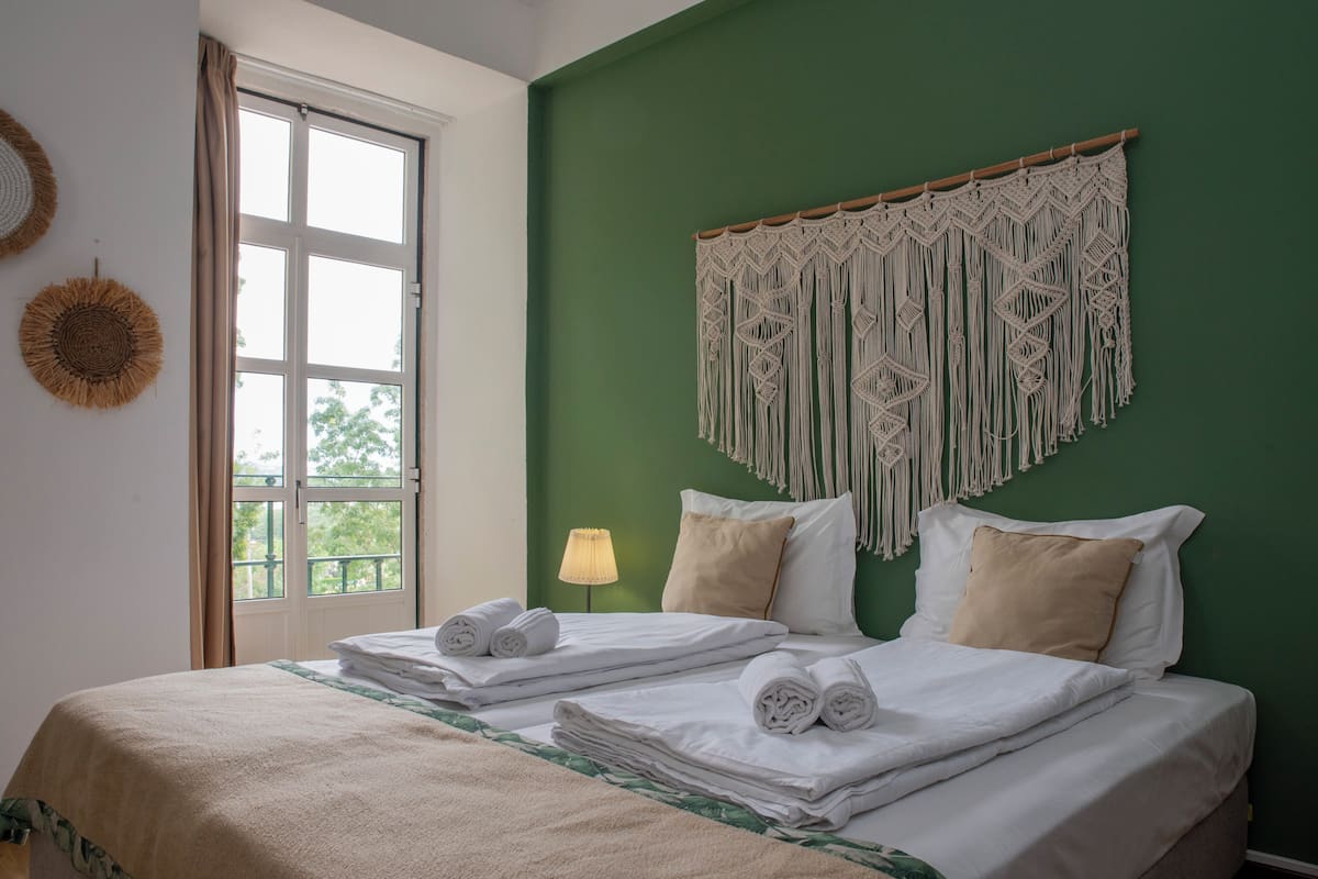 Een opgemaakt bed met witte lakens en twee opgerolde handdoeken, een macramé wanddecoratie en een raam met gordijnen.