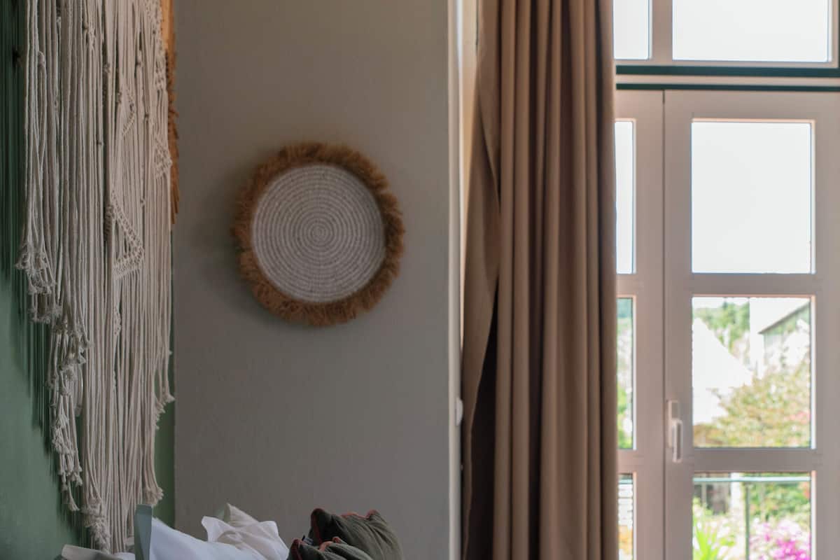 Een slaapkamer met een bed, kussens, een macramé wanddecoratie en uitzicht door een raam met gordijnen.