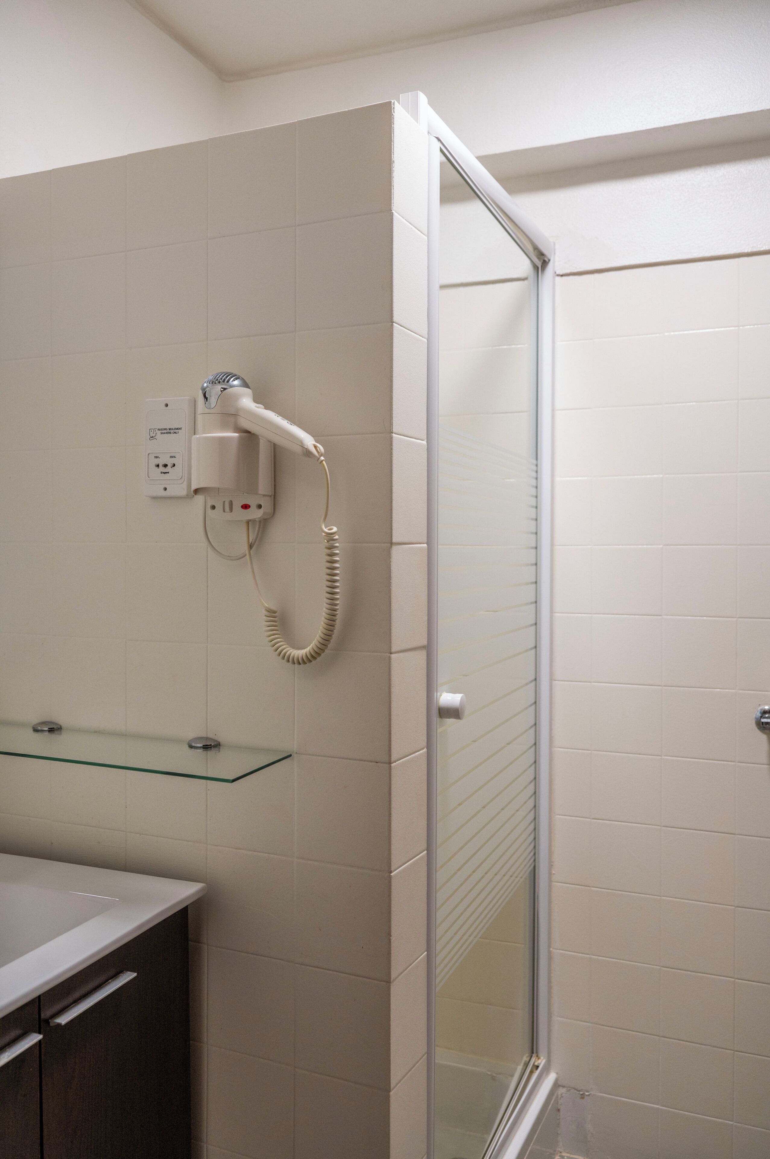 Un baño con cabina de ducha de vidrio, un secador de pelo blanco fijado a la pared y un estante de vidrio sobre un mueble de color oscuro.