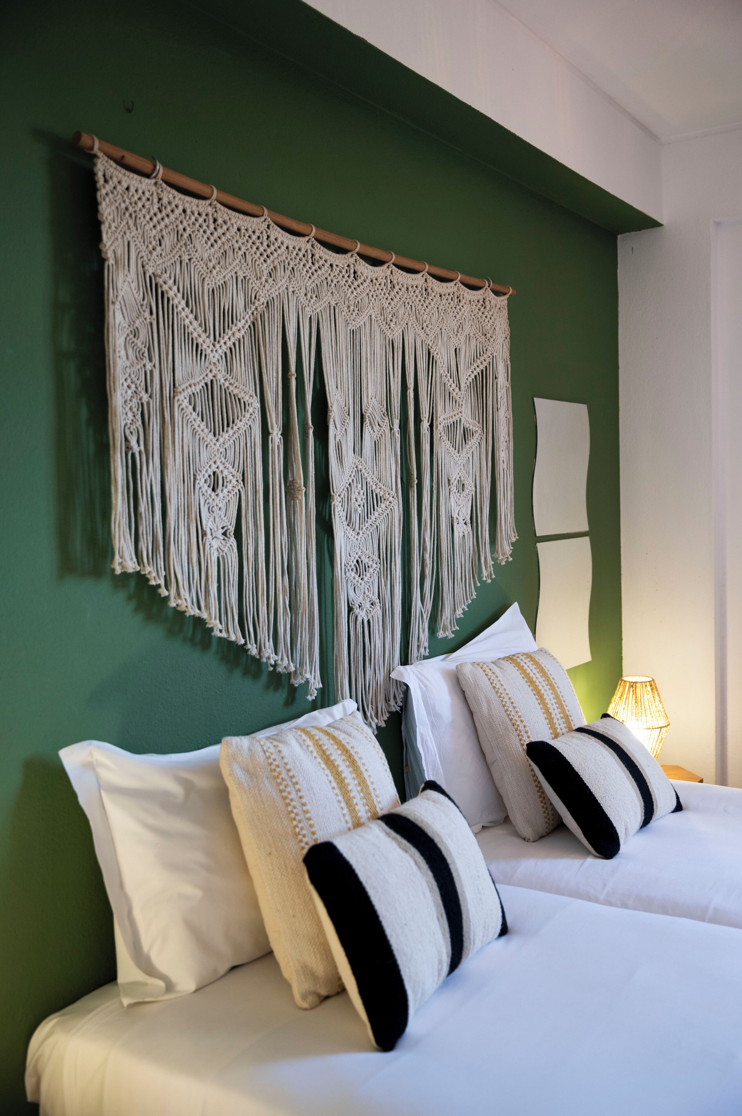 Una cama con ropa blanca y dos almohadas, un tapiz de macramé en la pared y una lámpara de noche.