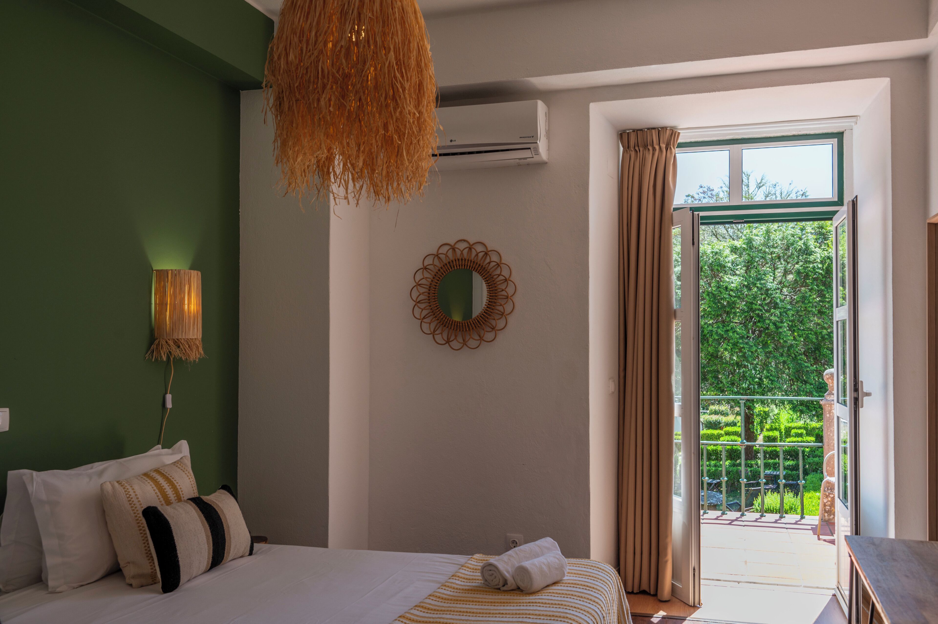Une chambre à coucher comprenant un lit, un luminaire suspendu, un appareil de climatisation, un miroir décoratif et une vue sur un jardin verdoyant par une porte ouverte.