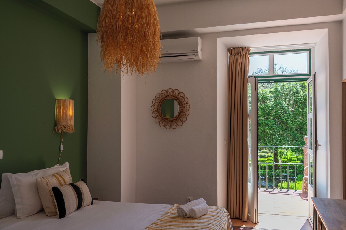 Un dormitorio con cama, lámpara colgante, equipo de aire acondicionado, espejo decorativo y vista a un jardín verde a través de una puerta abierta.