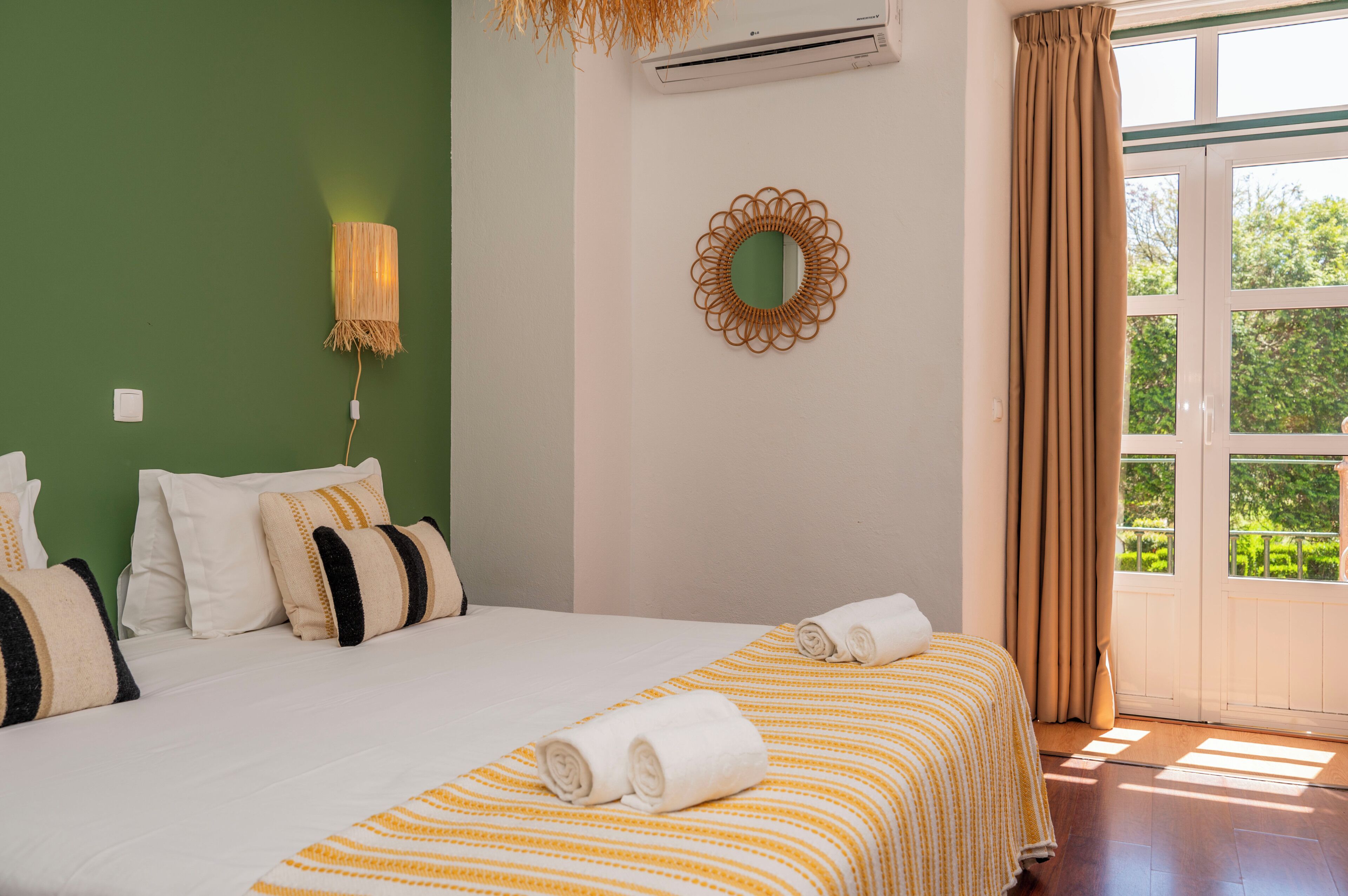 A hotel room with a bed, pillows, a wall-mounted air conditioner, a mirror, and a window with curtains.