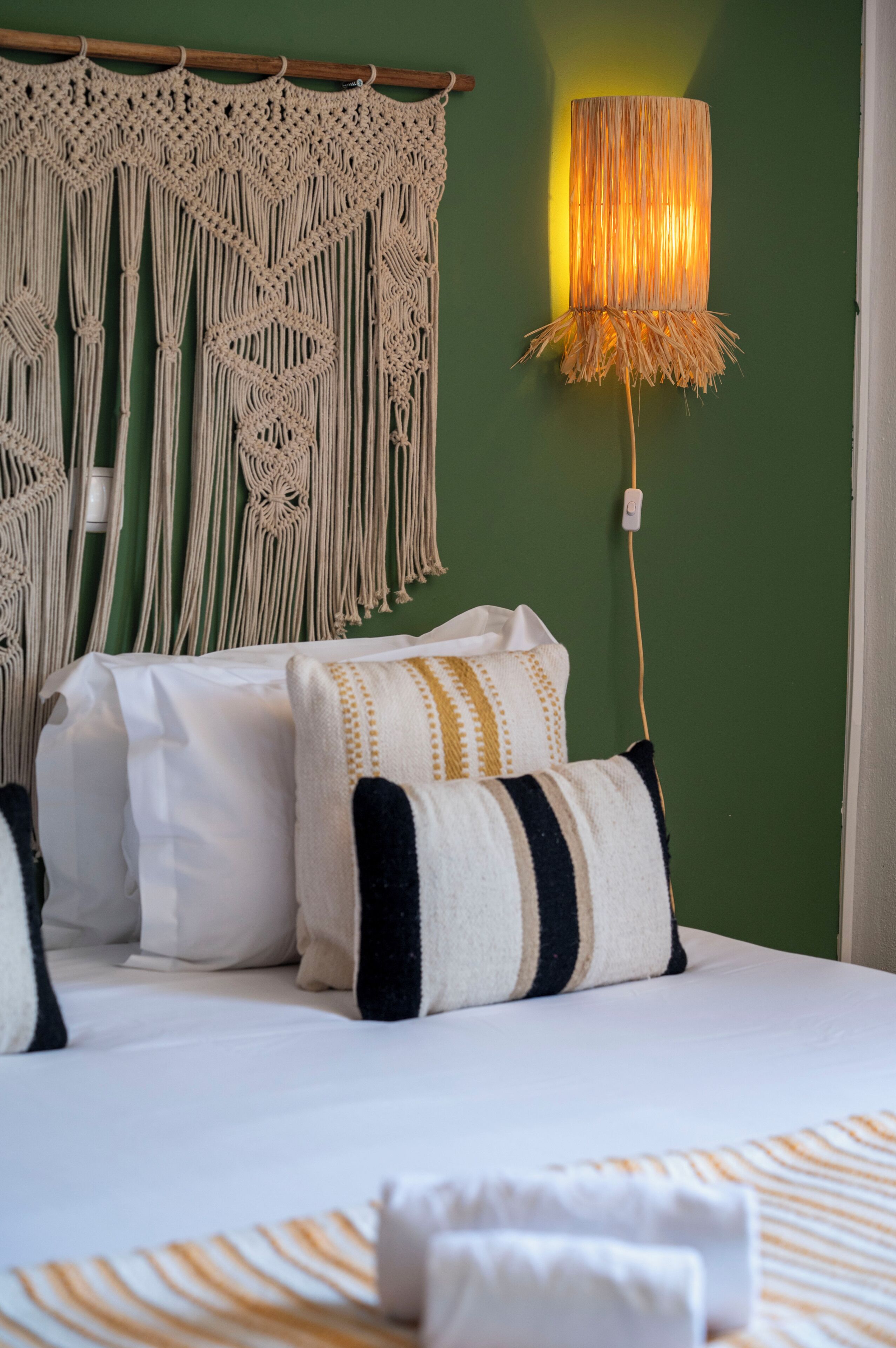 A bed with white bedding and decorative pillows, a macrame wall hanging, and a wall-mounted lamp.