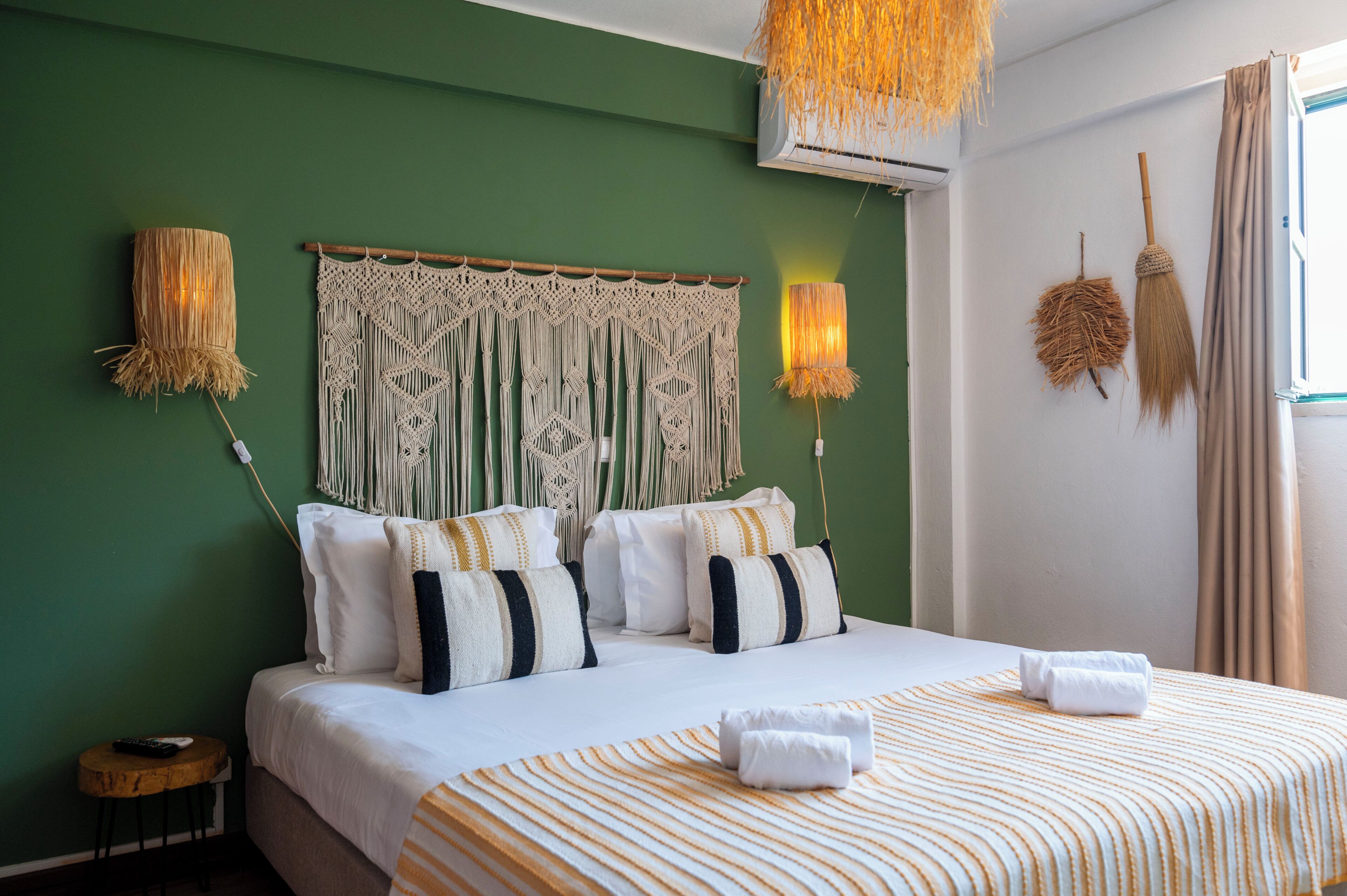 A hotel room with a bed, pillows, a macrame headboard, wall-mounted lamps, and a window with curtains.
