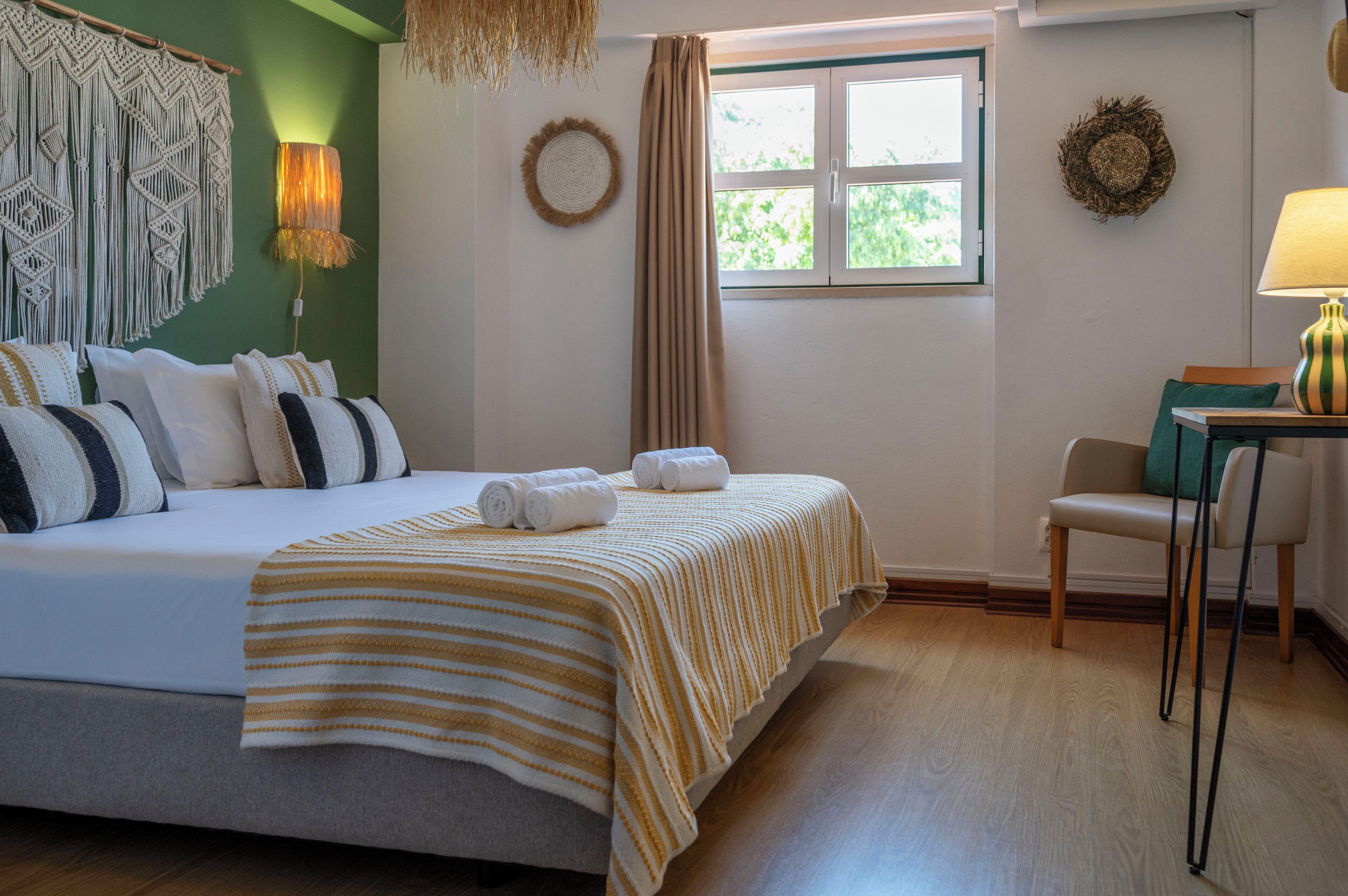 Habitación de hotel con cama, una silla, una mesa, una ventana y una pared decorada con un tapiz macramé y una obra de arte textil.