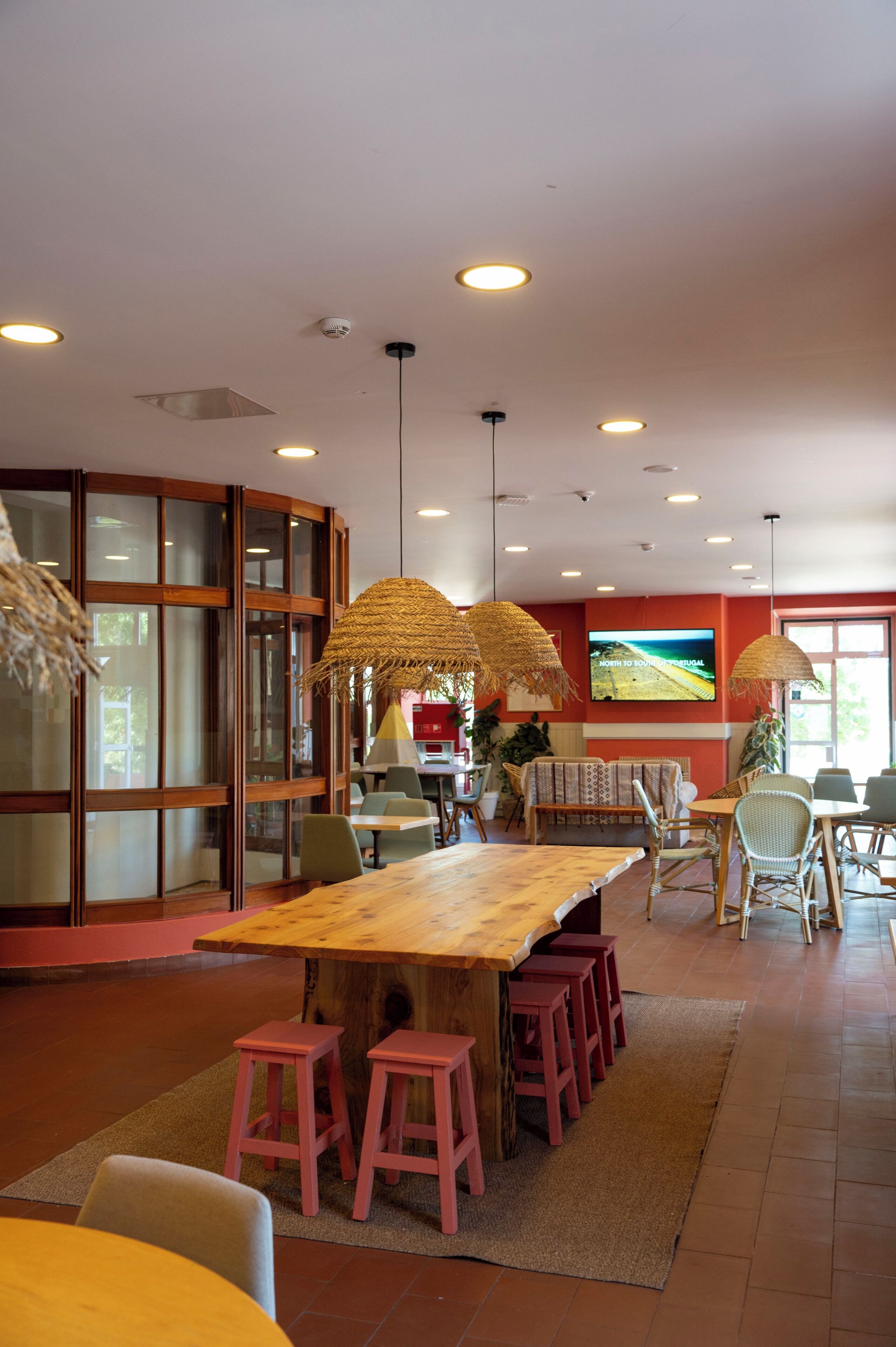 Un restaurante con una larga mesa de madera, taburetes rojos, lámparas de techo tejidas y una pantalla grande que muestra una vista panorámica.