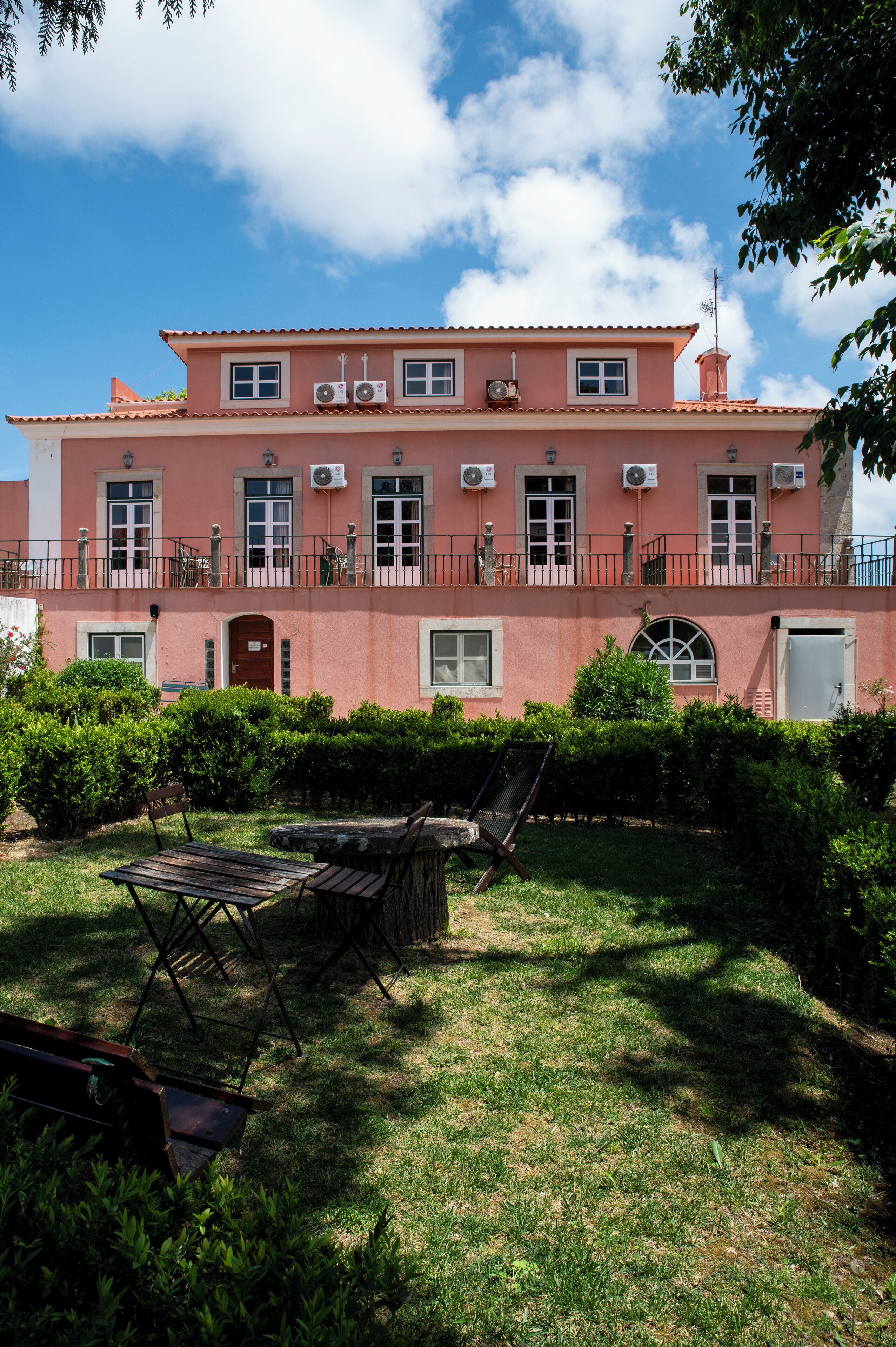 A pink building with multiple windows and balconies, surrounded by well-maintained hedges and a grassy lawn.