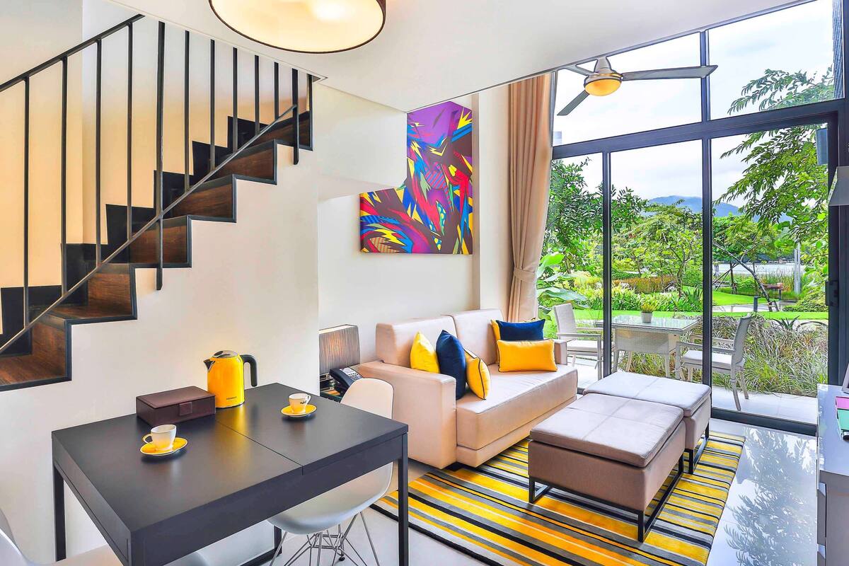 A modern living room with a staircase, a dining area, and large windows offering a view of greenery.