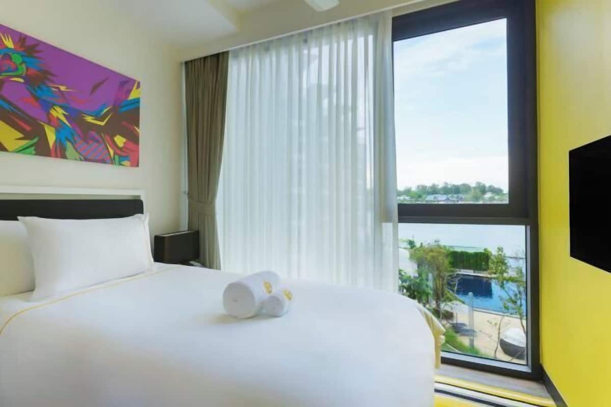 A hotel room with a bed, a large window, a painting on the wall, and a view of a pool and greenery.