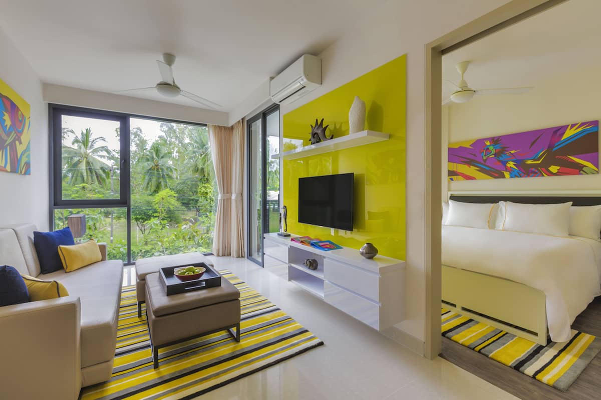 A modern hotel room with a large window, a flat-screen TV, a bed, and a bright yellow accent wall.