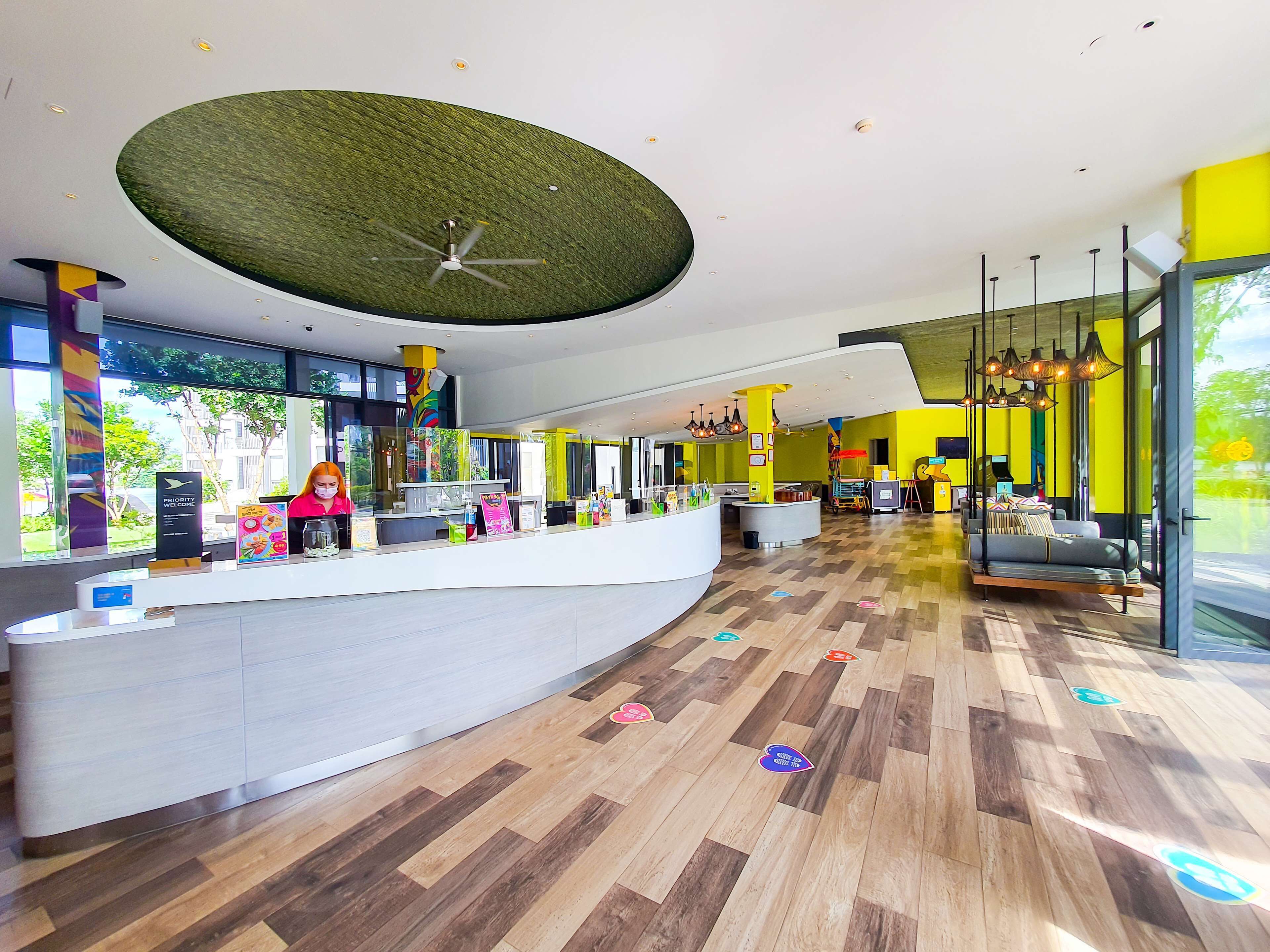A modern hotel lobby with a curved reception desk, a ceiling fan, and a large window offering a view of the outdoors.