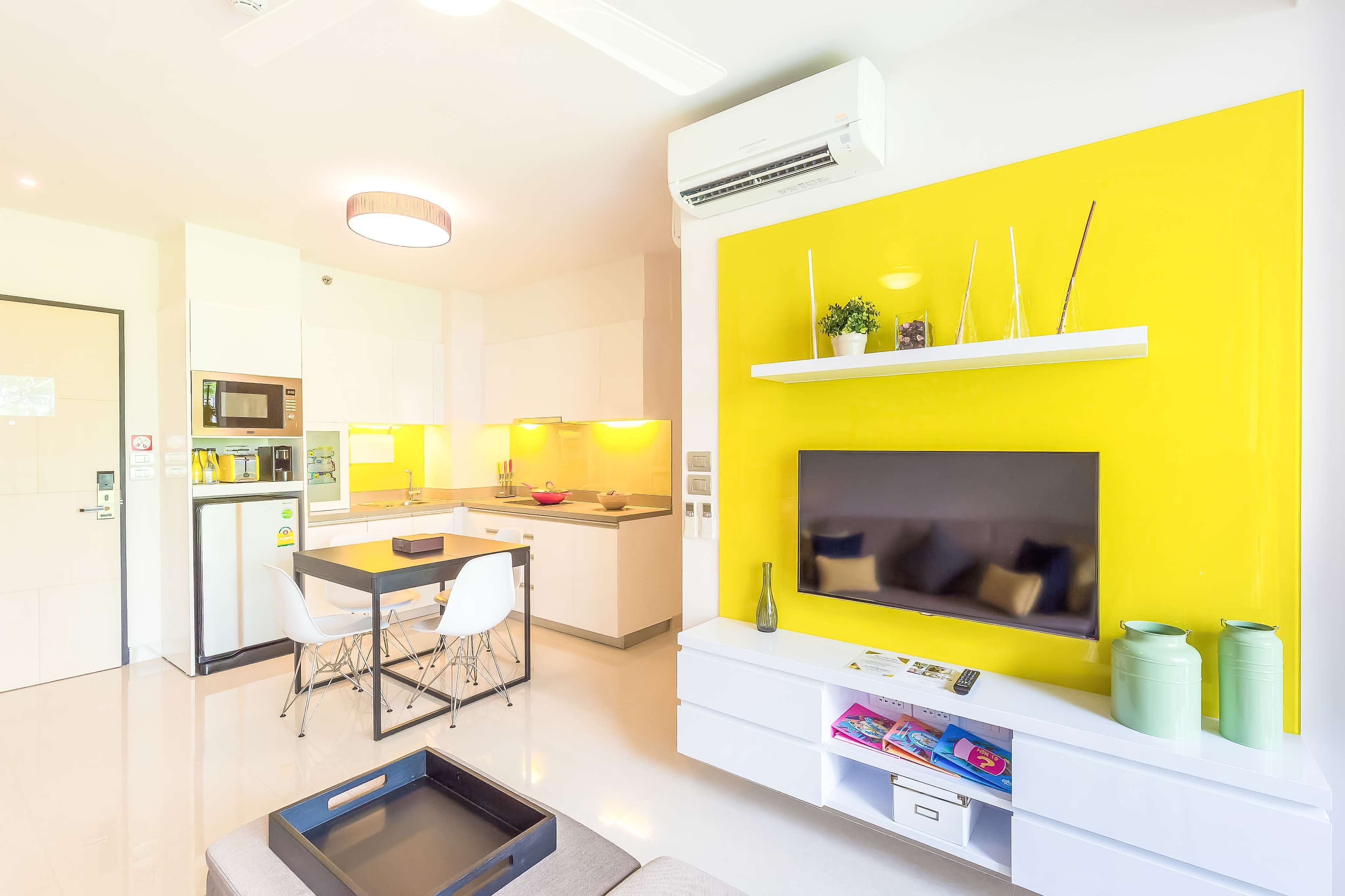 A modern kitchen with a dining area, a flat-screen TV mounted on a yellow accent wall, and a small seating area with a table and chairs.