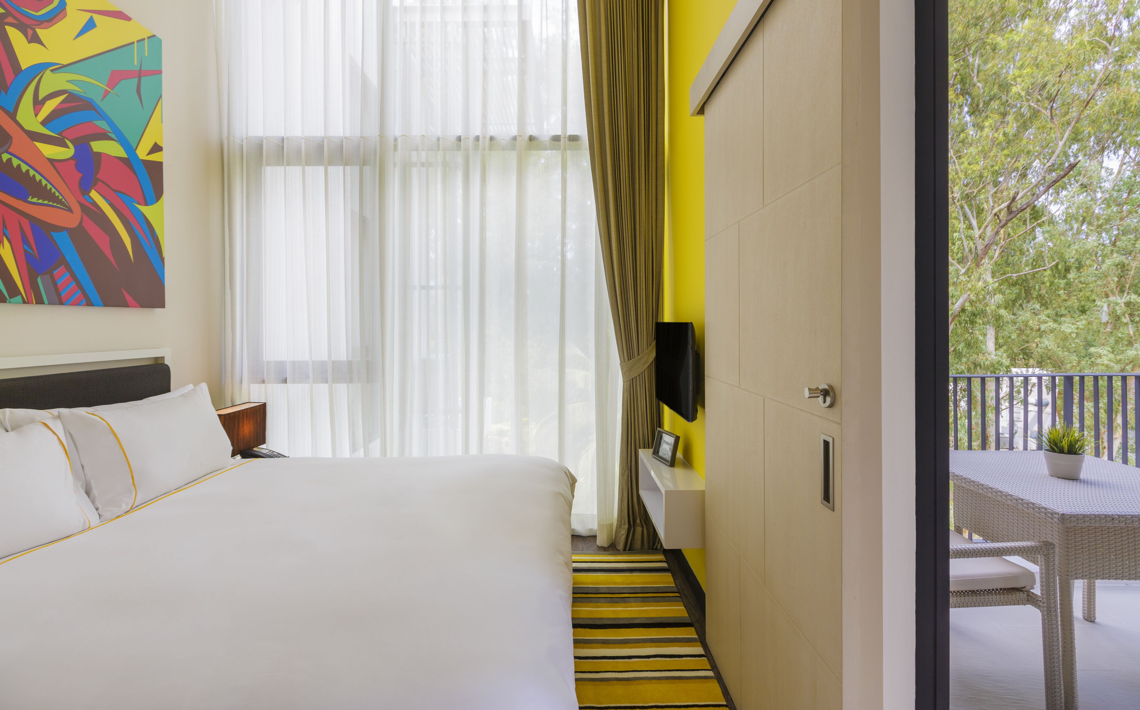 Una camera d'albergo con un letto, una televisione a parete, una finestra con tende leggere, una parete gialla e un balcone con tavolo e sedia.