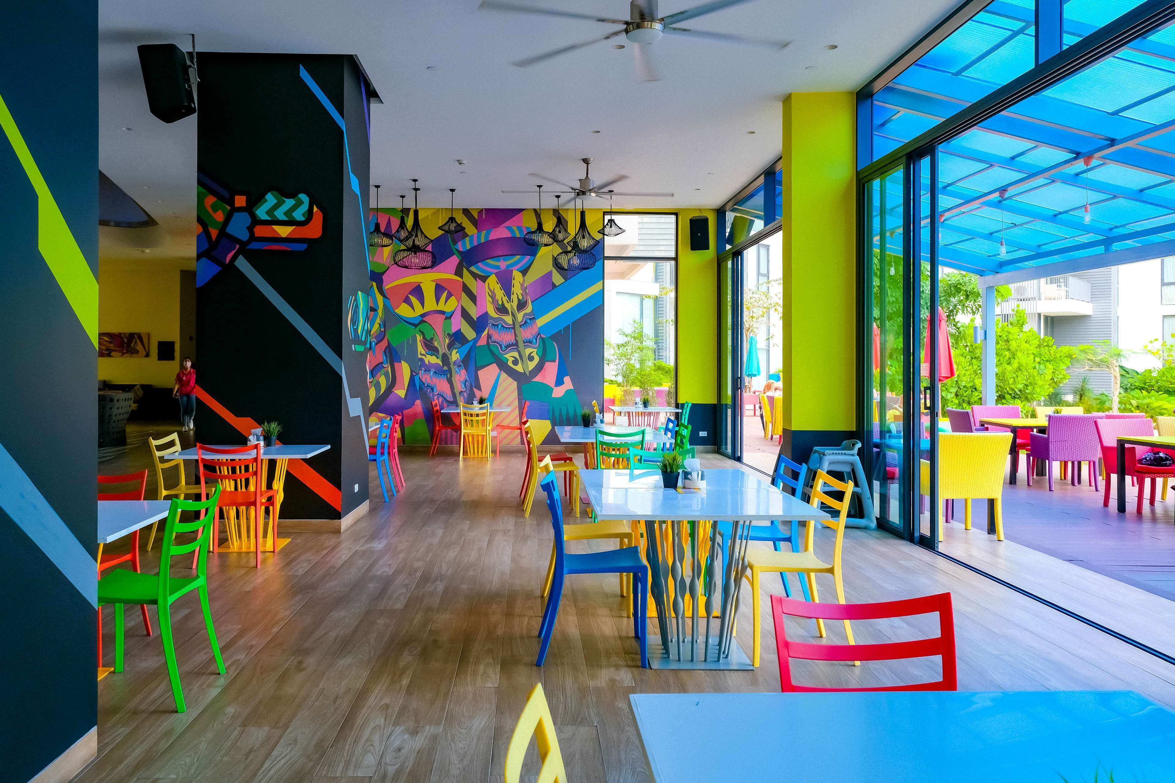 A vibrant indoor space with colorful chairs and tables, a ceiling fan, and a large window with a view of the outdoors.