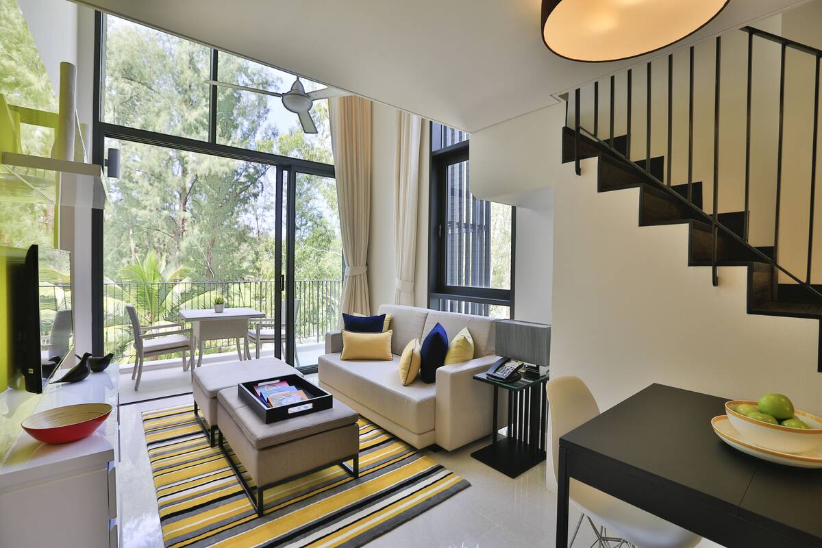 A modern living room with a sofa, coffee table, and dining area. There is a staircase and large windows with curtains.