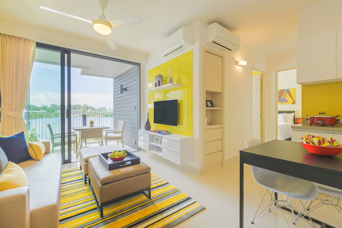 A modern living room with a sofa, ottoman, and a dining table with chairs. There is a TV on a white cabinet, and a balcony with a view.