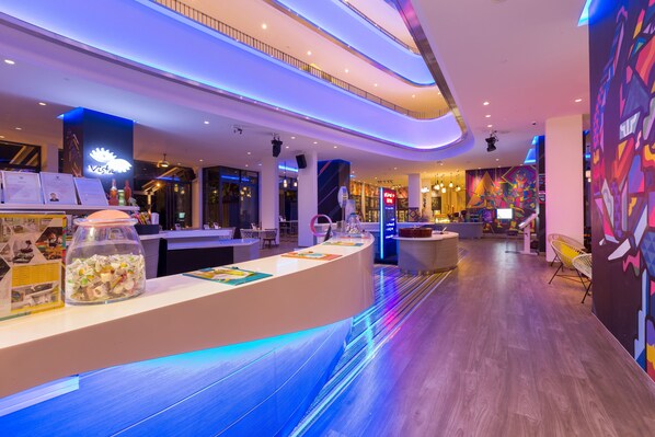 Eine moderne Hotelhalle mit einer gebogenen Rezeption, einer Glasvitrine mit Snacks und einem farbenfrohen Wandbild.